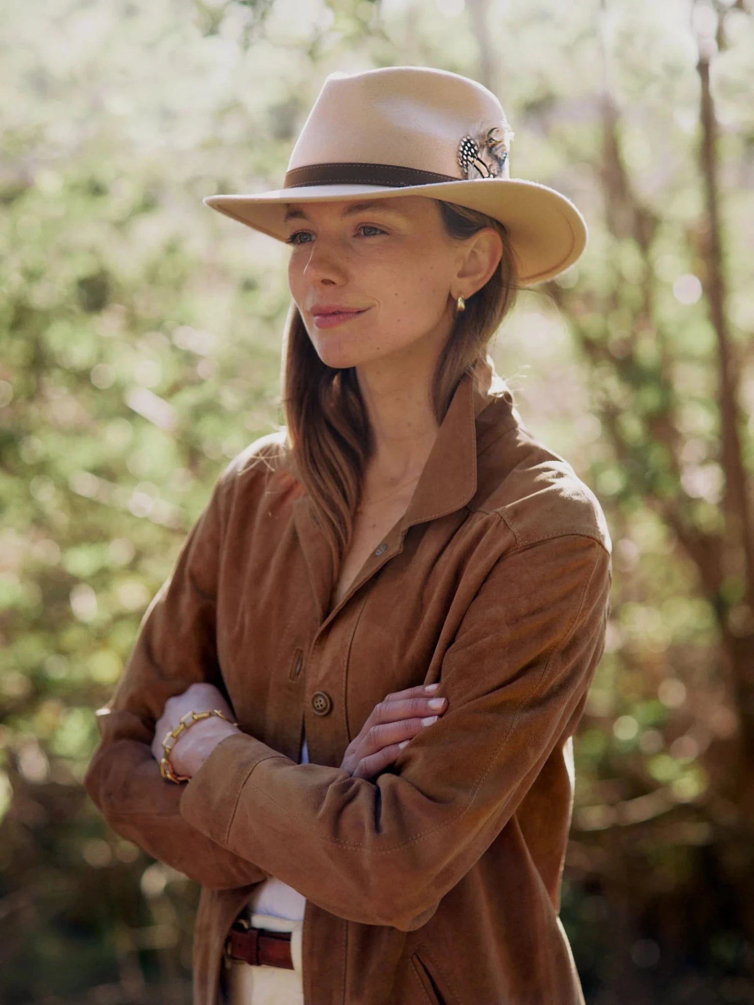A woman stands outdoors with her arms crossed, wearing a Hicks & Brown Auld Stock Gamebird Fedora in beige wool felt with a feather wrap and a brown suede jacket, surrounded by blurred greenery.