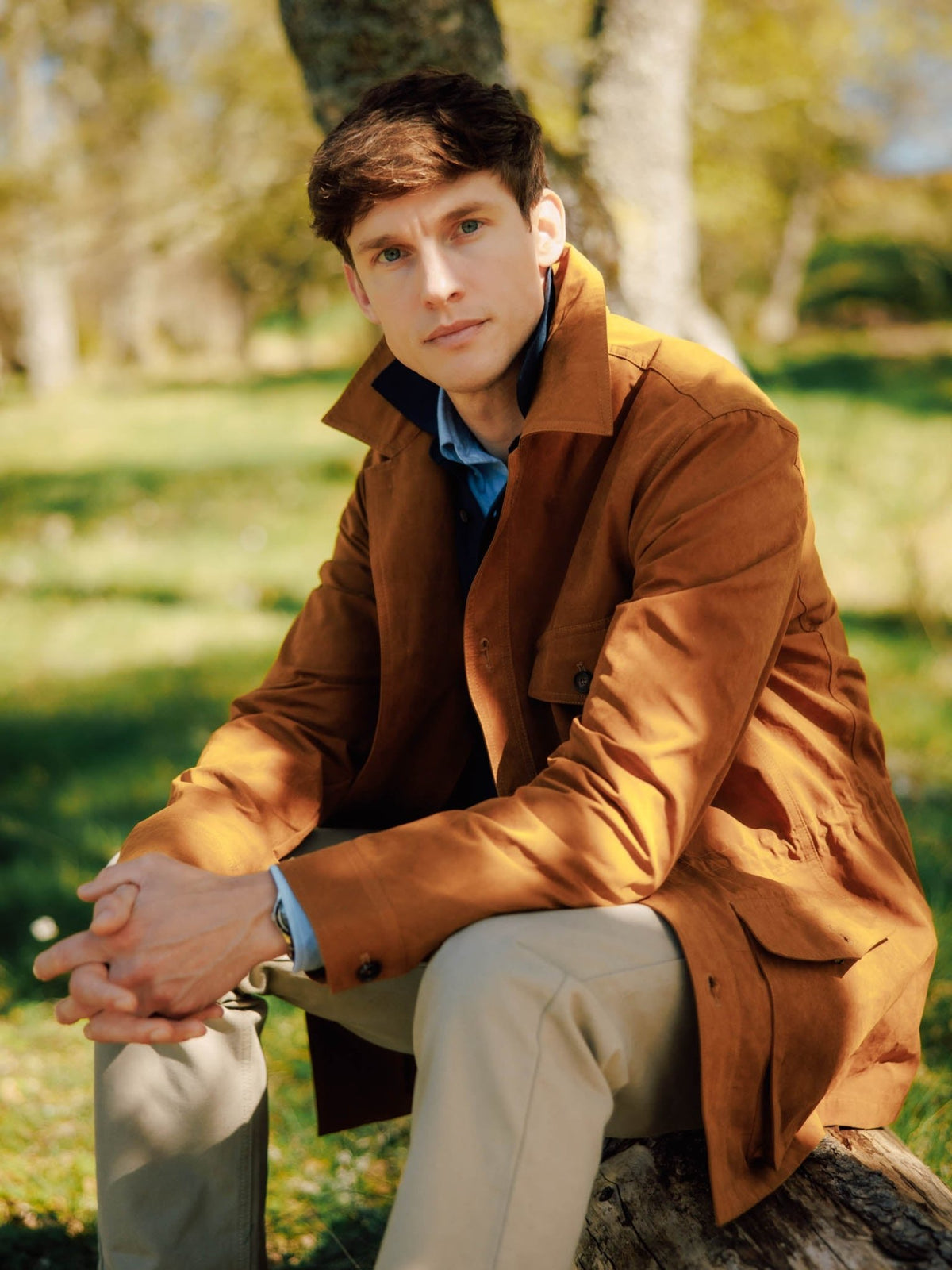 A man wearing a tan Italian Aigas Field Jacket by Campbell’s of Beauly and light-colored trousers sits on a fallen log outdoors.