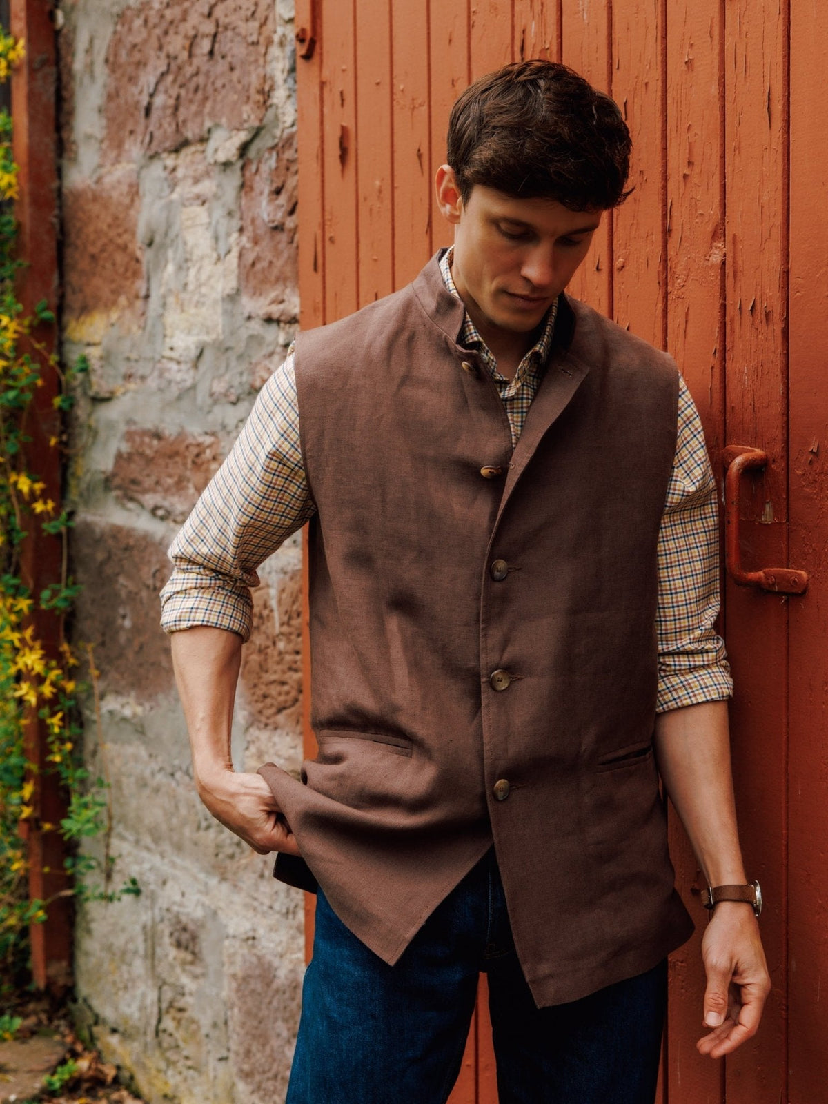 A man wears a brown linen Auld Stock Linen Nehru Gilet over a yellow and blue plaid long-sleeved shirt and blue jeans.