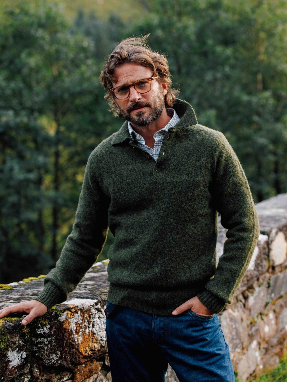 A man wearing a dark green Brushed Shetland Rugby Jumper over a collared shirt, standing by a stone wall outdoors.
