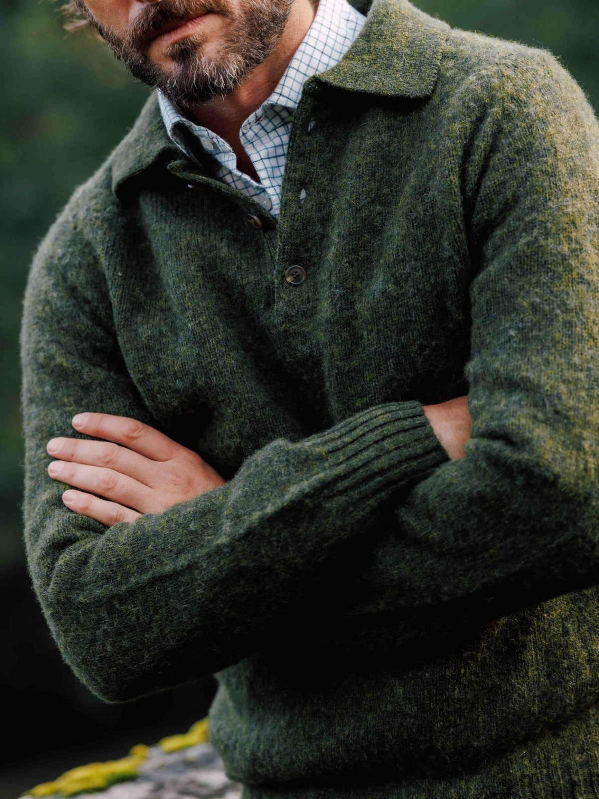 A man with his arms crossed wearing a dark green Brushed Shetland Rugby Jumper over a light-colored plaid shirt.