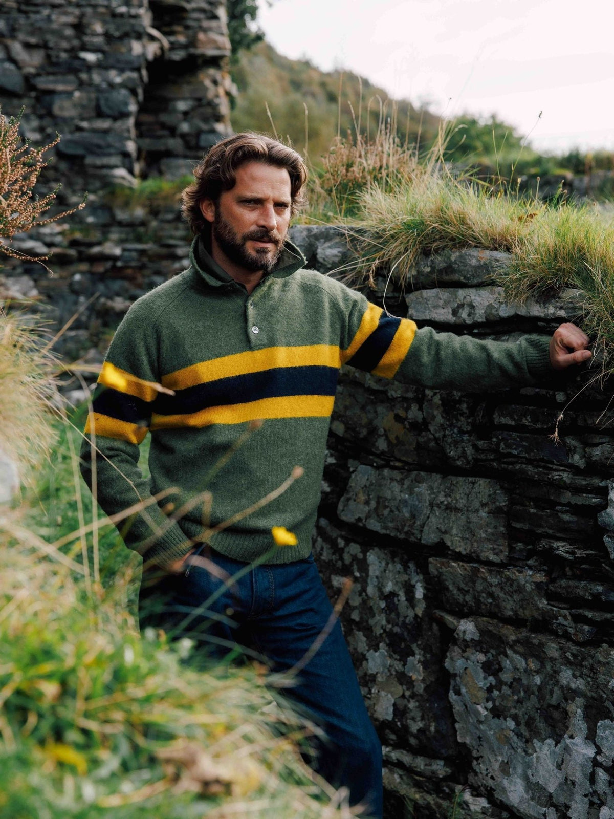 A man wearing a Campbell’s of Beauly Brushed Shetland Stripe Rugby, a green wool sweater with two horizontal yellow and dark navy stripes across the chest and sleeves.