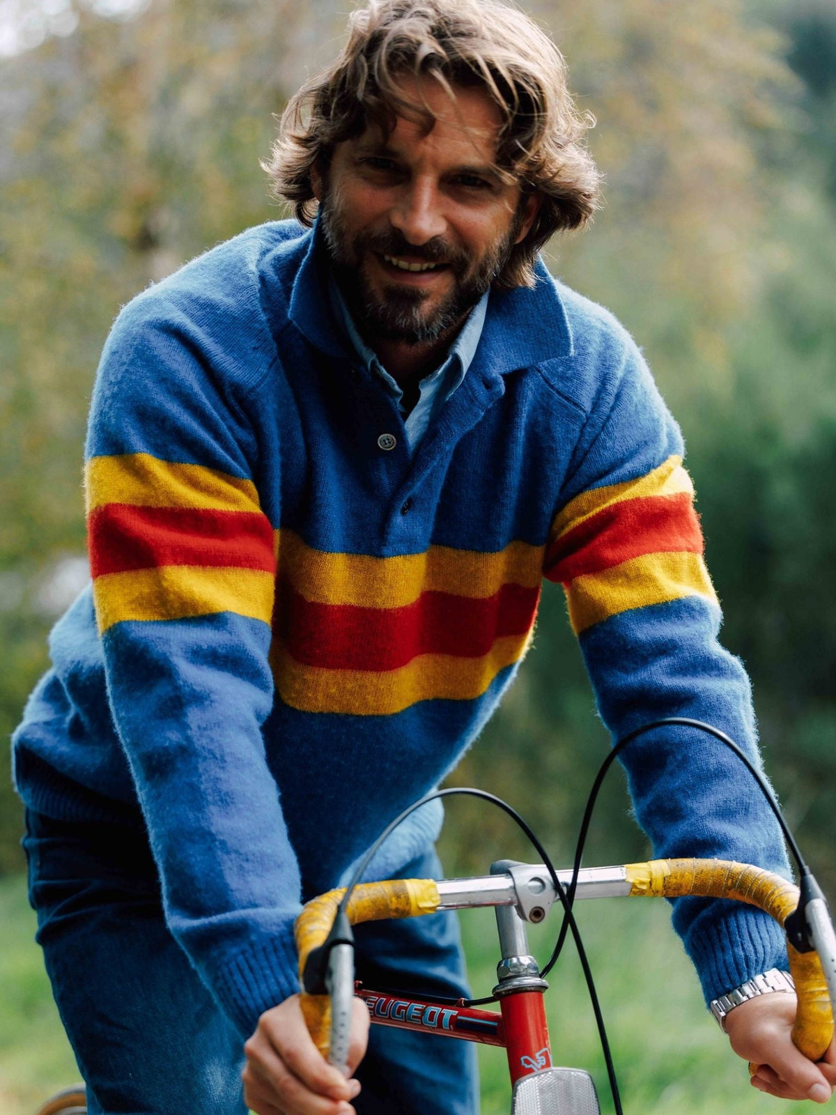 A man smiling while riding a bicycle, wearing a Brushed Shetland Stripe Rugby with wide yellow and red horizontal stripes across the chest.