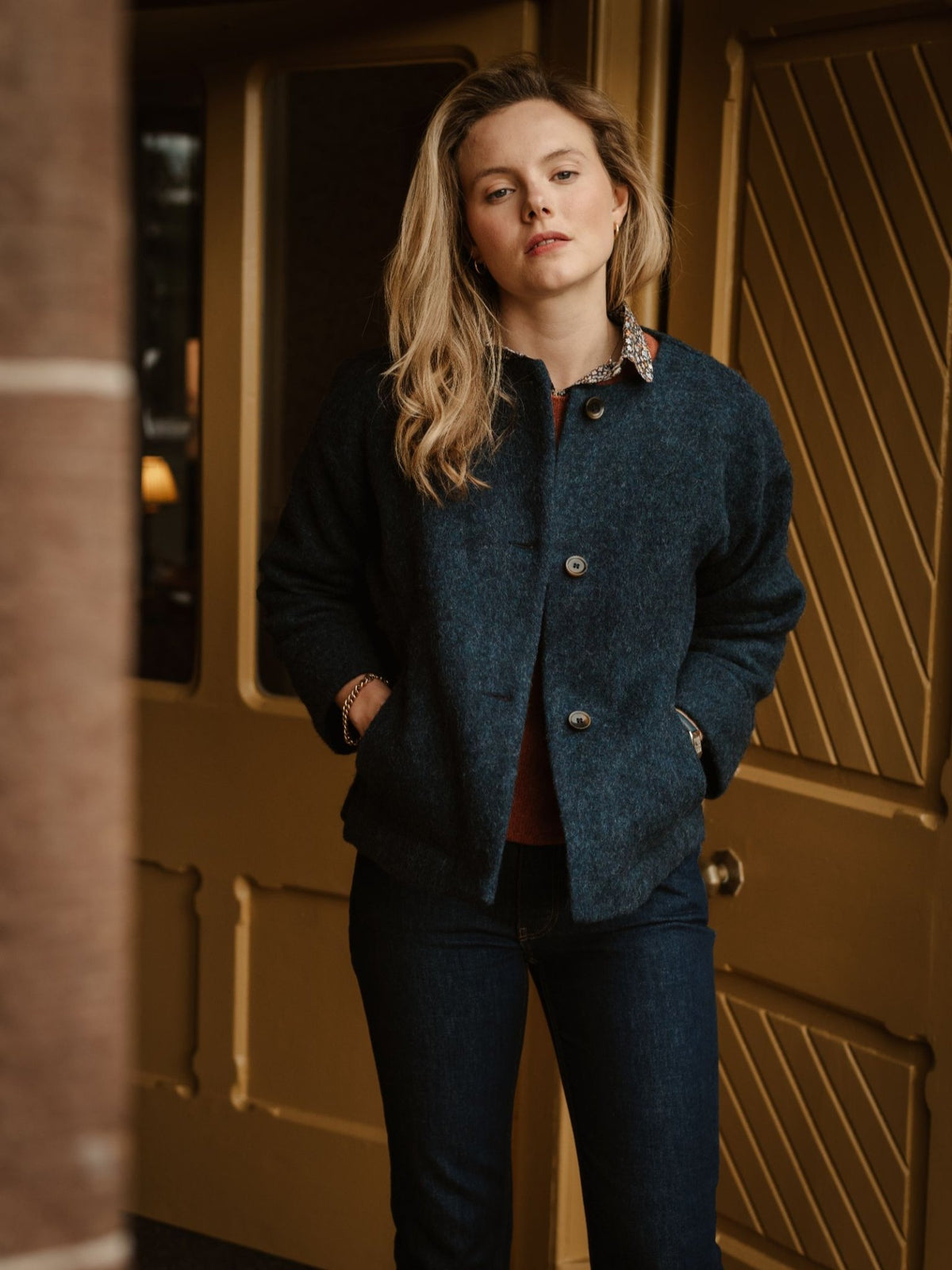 A woman with long blonde hair stands indoors, wearing the Campbell’s of Beauly Brushed Wool Jacket in navy and jeans, with her hands in her pockets, looking directly at the camera. Yellow paneled doors are in the background.