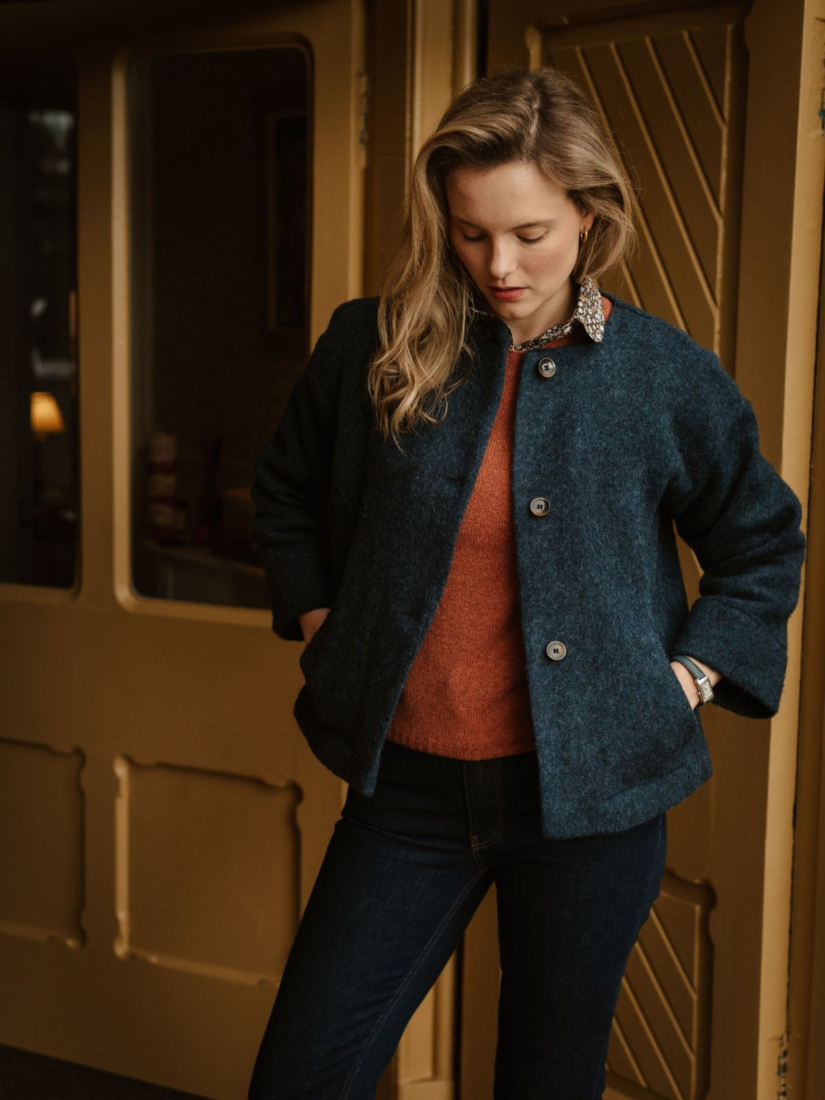 A woman with long blonde hair stands indoors, looking down. She wears the Brushed Wool Jacket by Campbell’s of Beauly—crafted from British boiled wool in dark blue—over an orange sweater and dark jeans, her hands tucked in her pockets. Yellow doors are visible in the background.