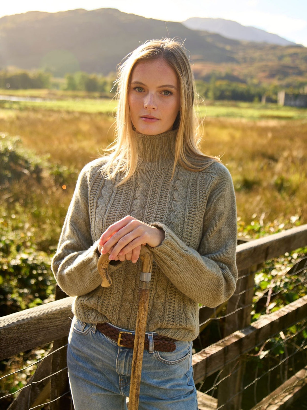 A woman wearing a supersoft wool Cable Roll Neck Jumper stands by a wooden fence holding a walking stick.
