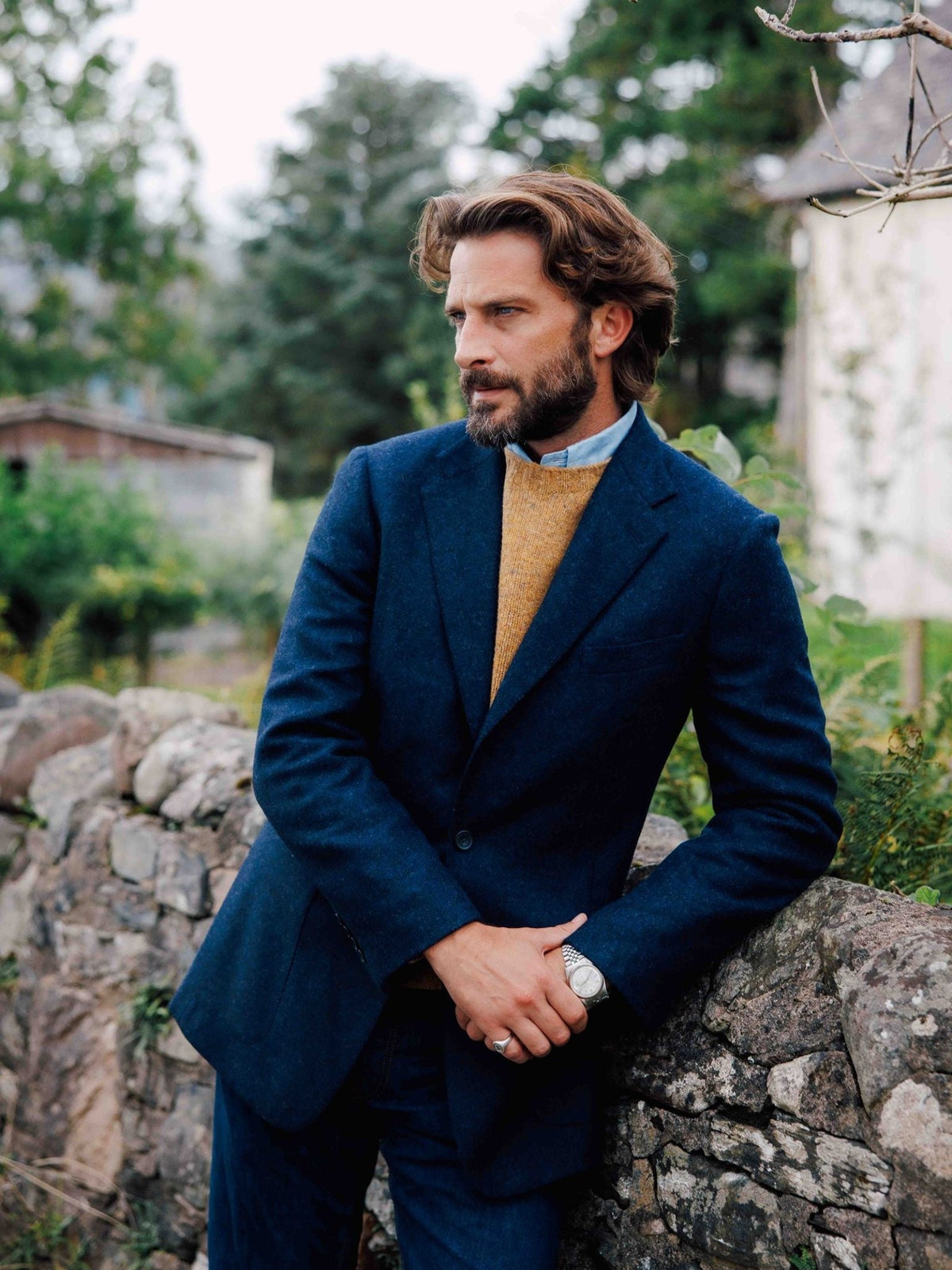 A man wearing a blue Abraham Moon Campbell’s House Jacket over a gold sweater and light blue shirt, leaning against a stone wall.