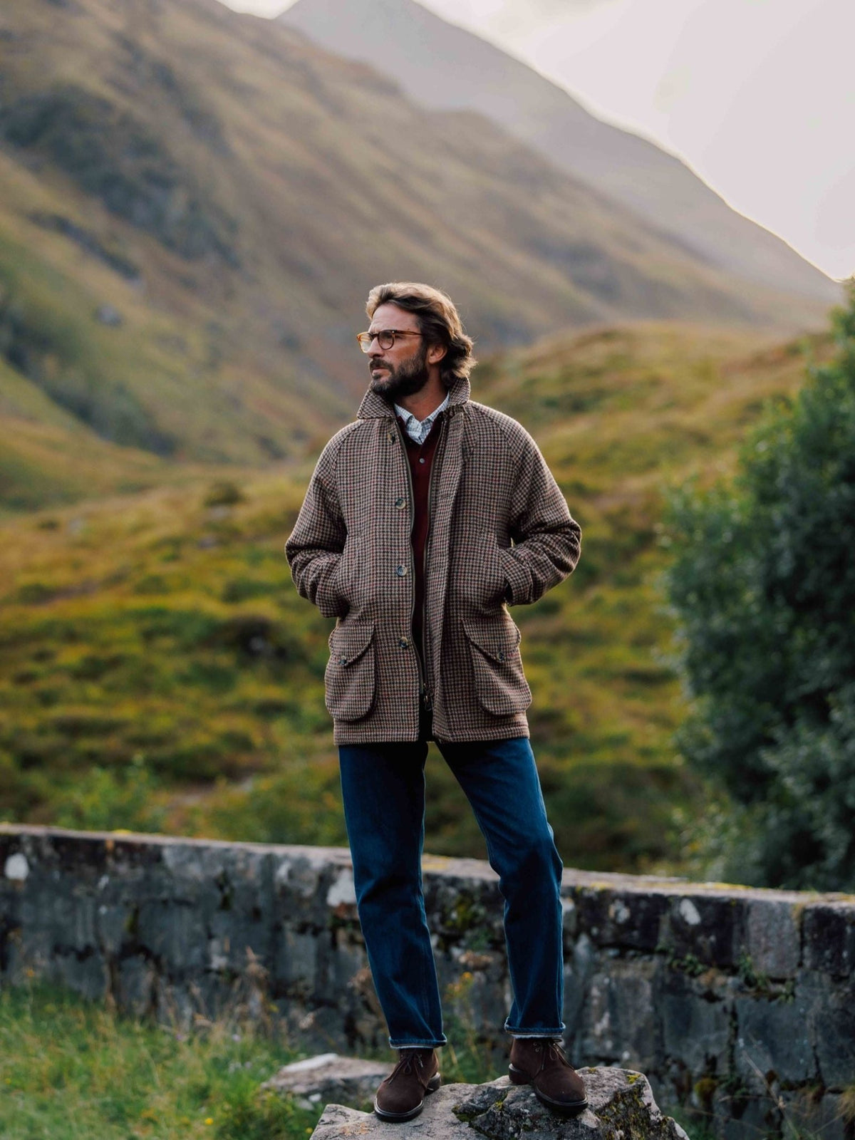 A man wearing a Campbell’s of Beauly Cannich Coat, burgundy sweater, and dark jeans stands on a stone wall against a mountainous landscape.
