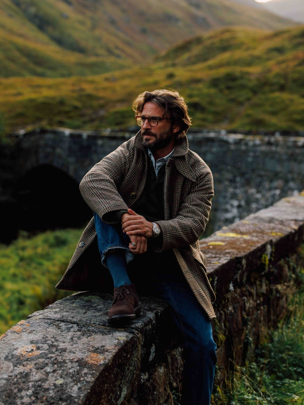 A bearded man wearing glasses and a Campbell’s of Beauly Cannich Coat sits on a stone bridge wall, dressed in blue jeans and brown suede shoes.