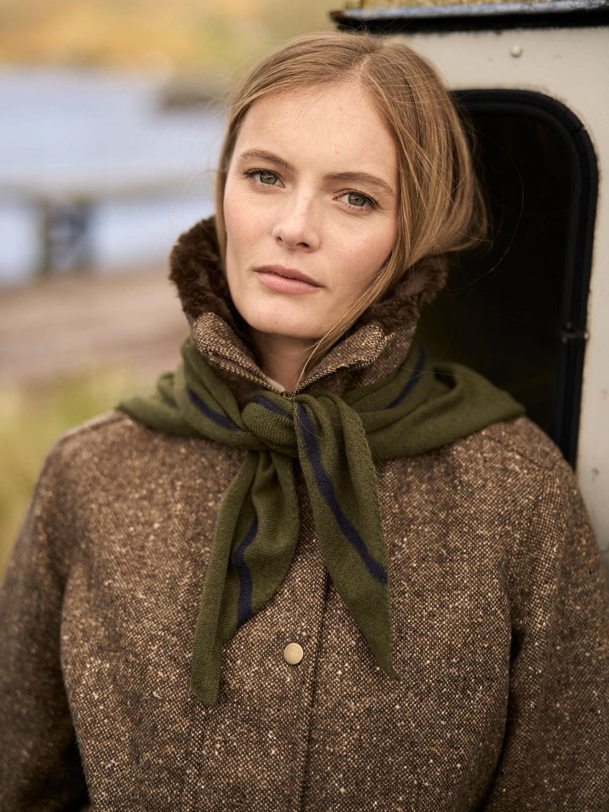 A woman wears an olive green and dark blue striped Campbell’s of Beauly Cashmere Neckerchief tied around her neck over a tweed jacket.