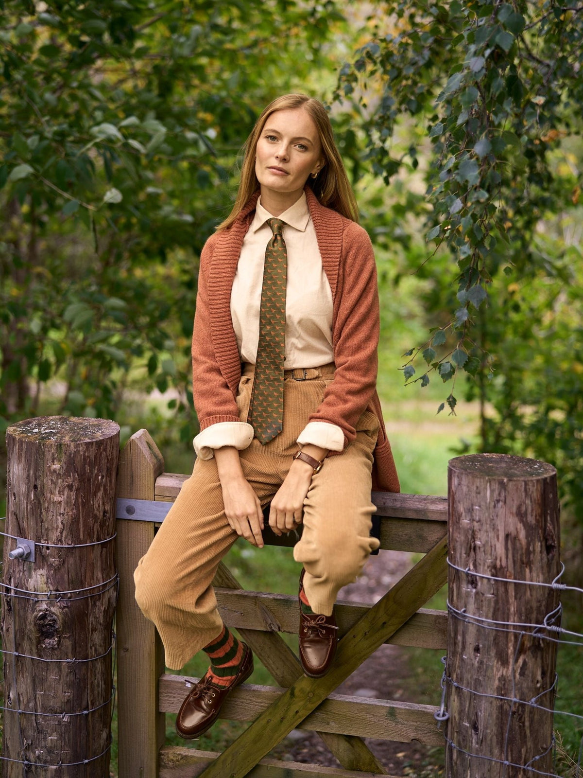 A woman wearing a rust-colored Campbell’s of Beauly Cashmere Shawl Cardigan over a tan shirt, patterned tie, and corduroy trousers sits on a wooden gate.