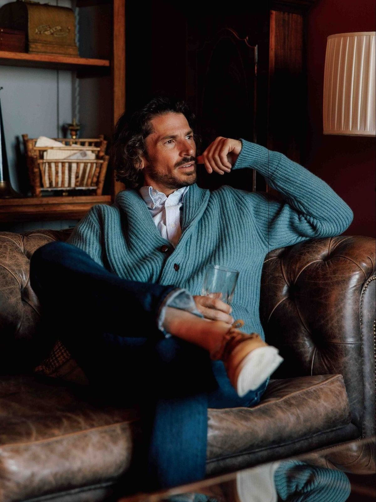 A man with wavy hair sits on a brown leather couch in a cozy, warmly lit room, wearing jeans and the Cashmere Shawl Collar Cardigan from Campbell’s of Beauly. He holds a glass and gazes thoughtfully to the side.