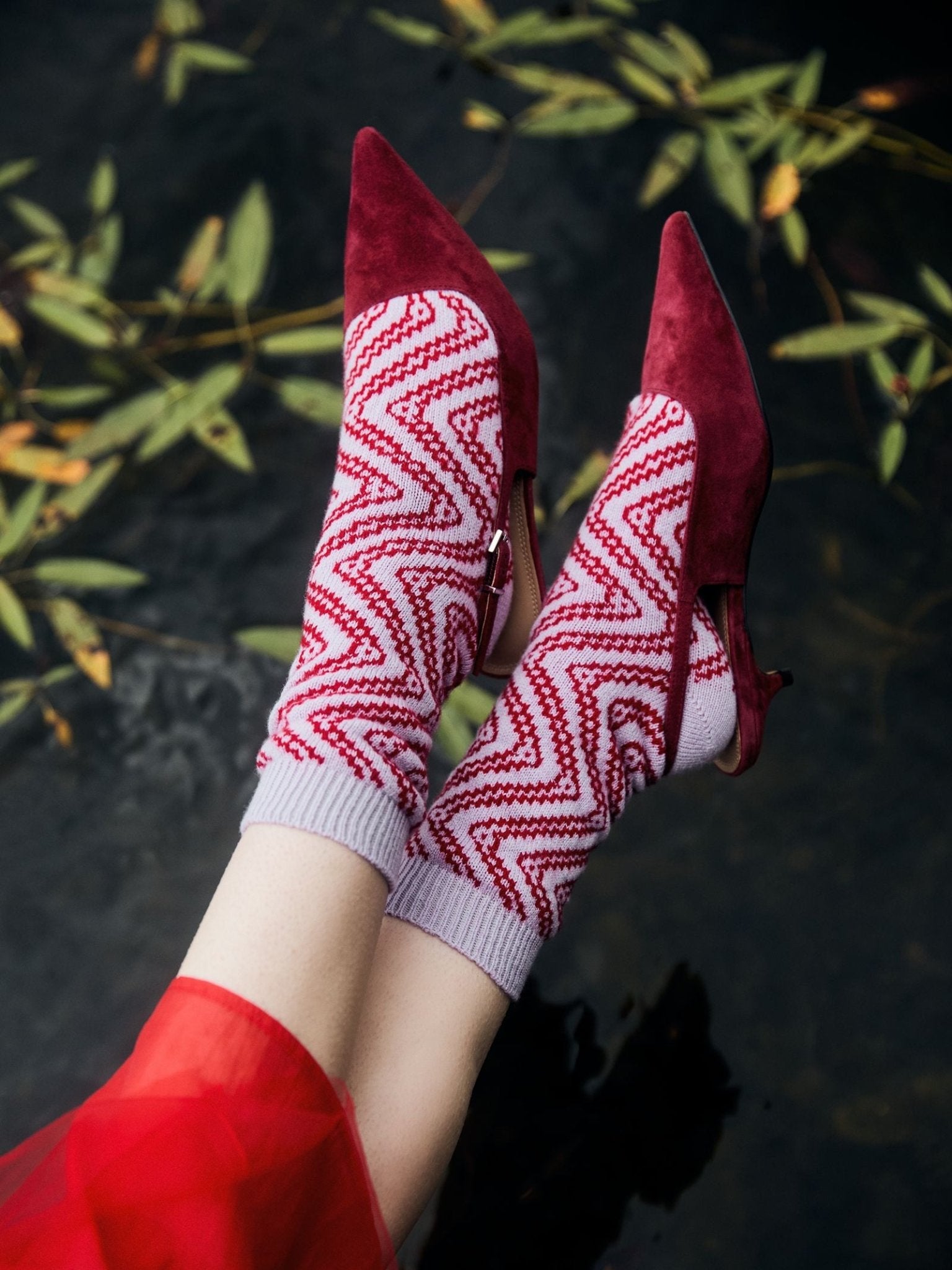 A person wears Campbell’s of Beauly Cashmere Swirly Socks—luxuriously warm with a red and white zigzag pattern—paired with red pointed-toe heels, legs crossed above dark water scattered with floating green leaves.