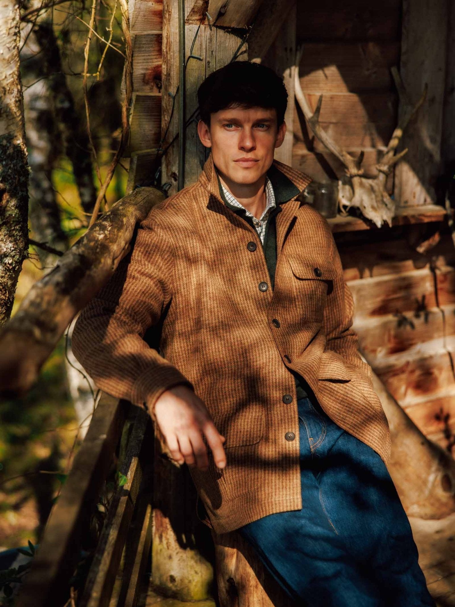 Wearing the Campbell’s of Beauly Casual Shacket and blue jeans, a man leans against a rustic wooden railing, sunlight and shadows playing over the scene. The wooden walls and antlers in the background enhance the relaxed sophistication of his look, with his effortless tailoring standing out in this casual setting.