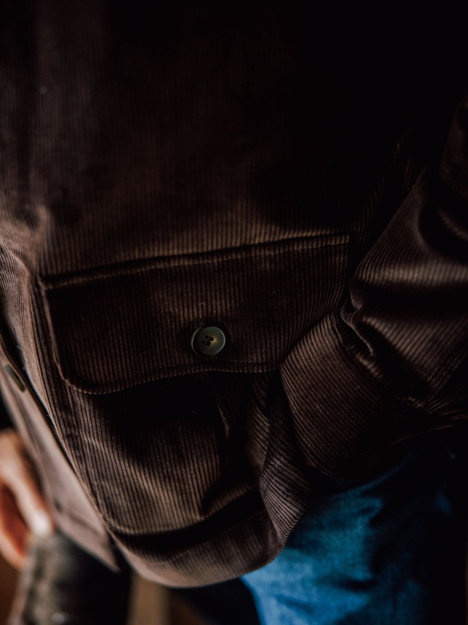 Close-up of the pocket on the Campbell’s of Beauly Casual Shacket in brown cotton corduroy, featuring a button detail. The relaxed style is captured with a partial view of a blue pant leg and a hand at the side.