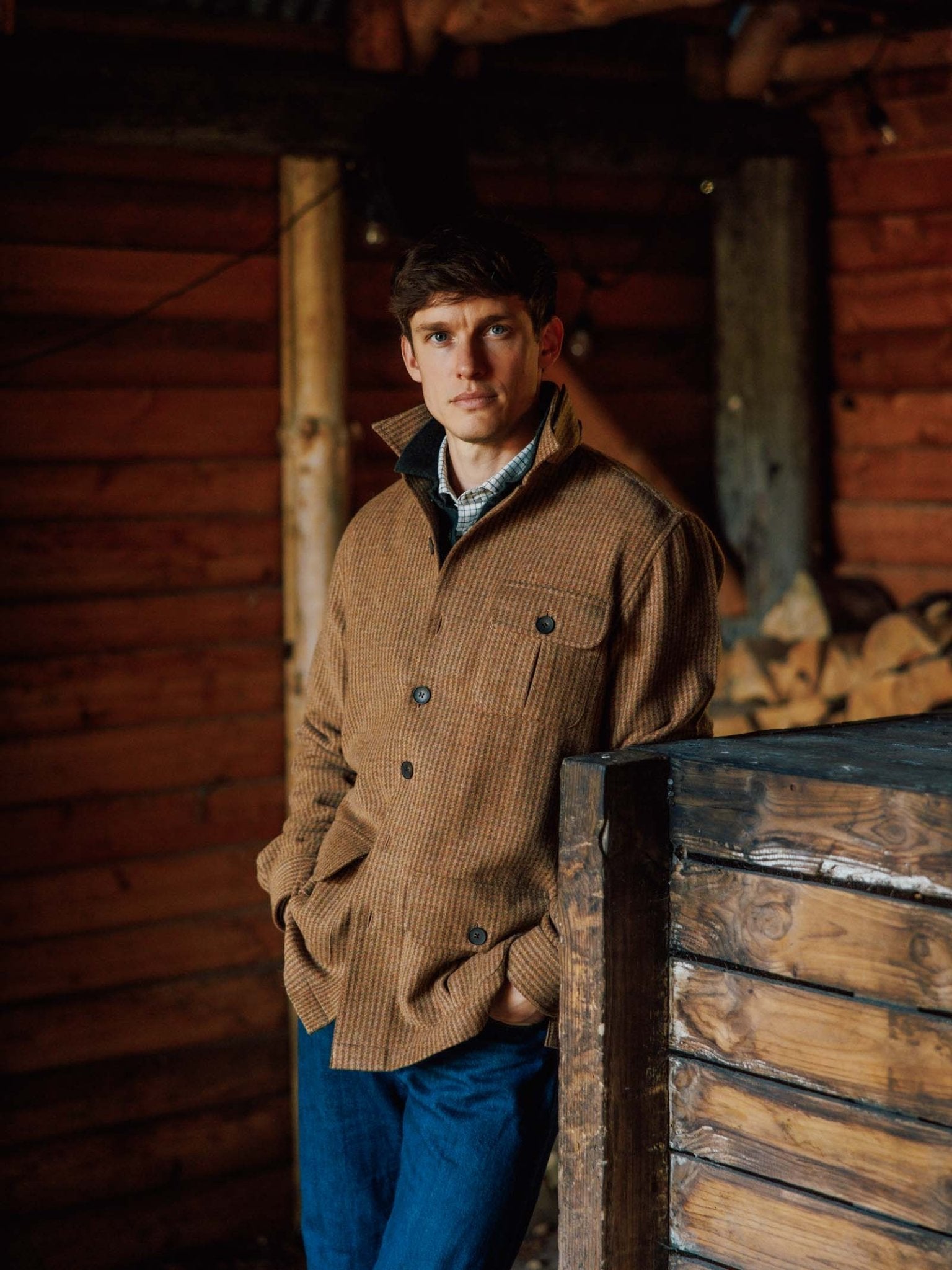 A man wearing a brown checked Casual Shacket from Campbell’s of Beauly and blue jeans stands indoors against a wooden wall, with stacked firewood in the background, highlighting relaxed tailoring inspired by classic British tweed.