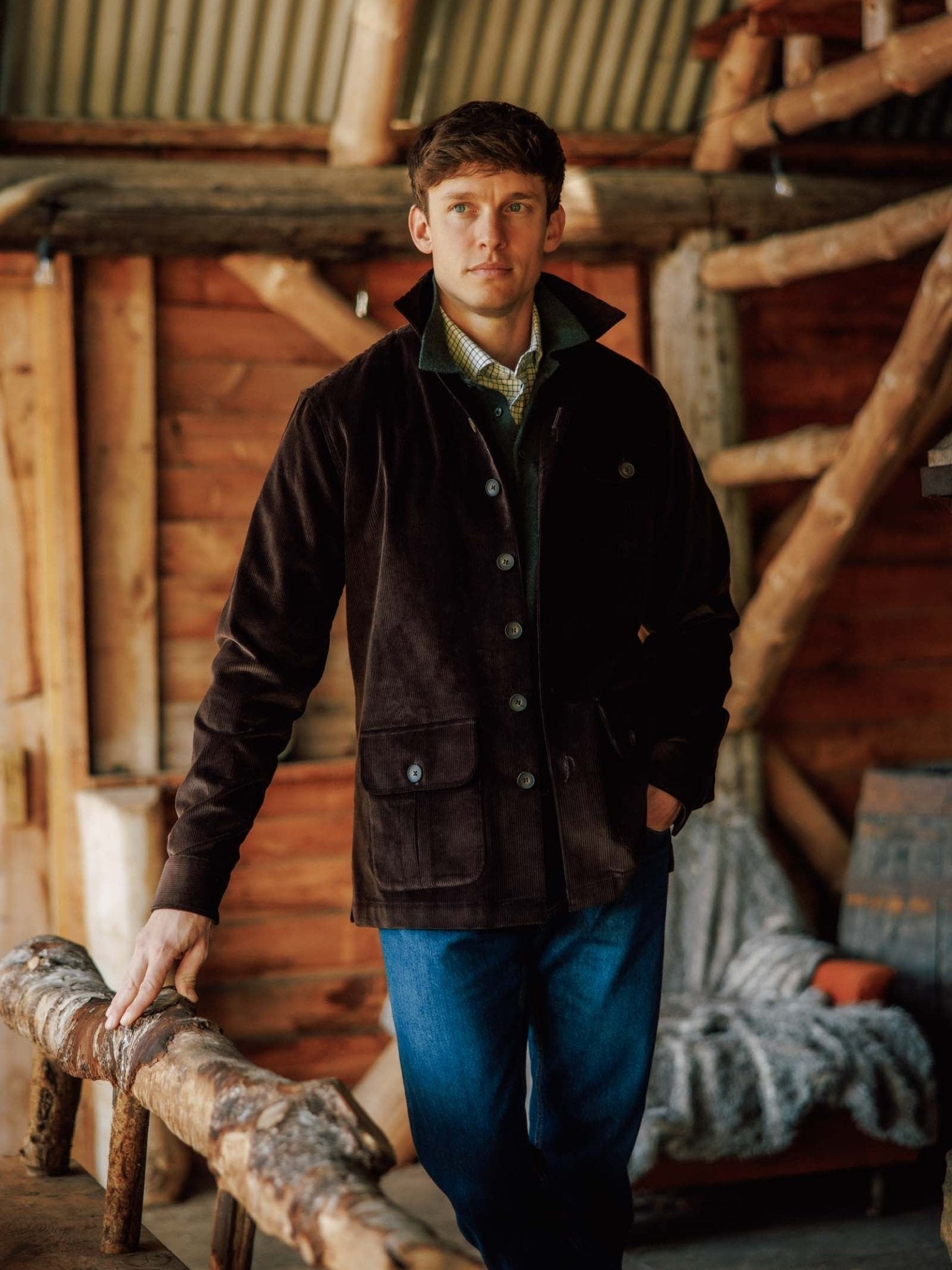 A man wearing the Campbell’s of Beauly Casual Shacket and blue jeans stands indoors by a rustic wooden railing, with wooden beams and cozy blankets in the background.