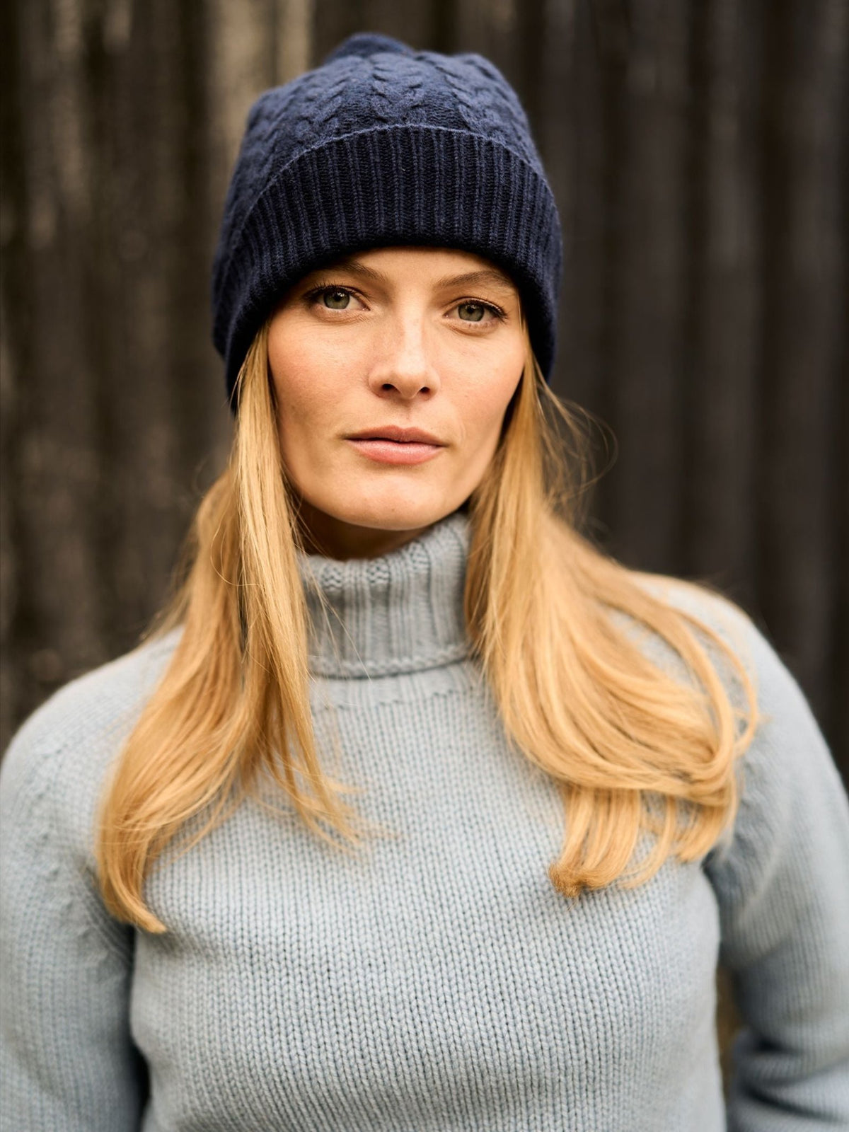 A woman wearing a dark blue Campbell’s of Beauly Chunky Cable Cashmere Hat with Pom Pom and a light blue turtleneck sweater.