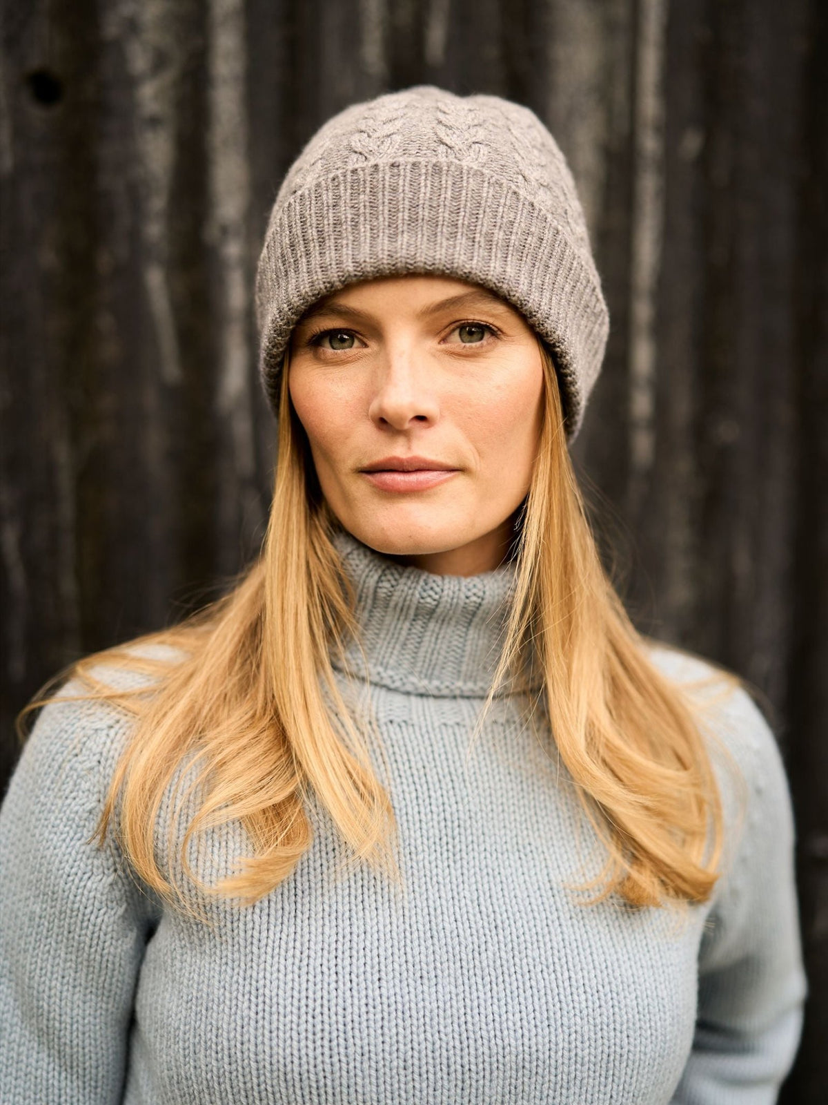 A woman wearing a light grey Chunky Cable Cashmere Hat with Pom Pom and a light blue turtleneck sweater.
