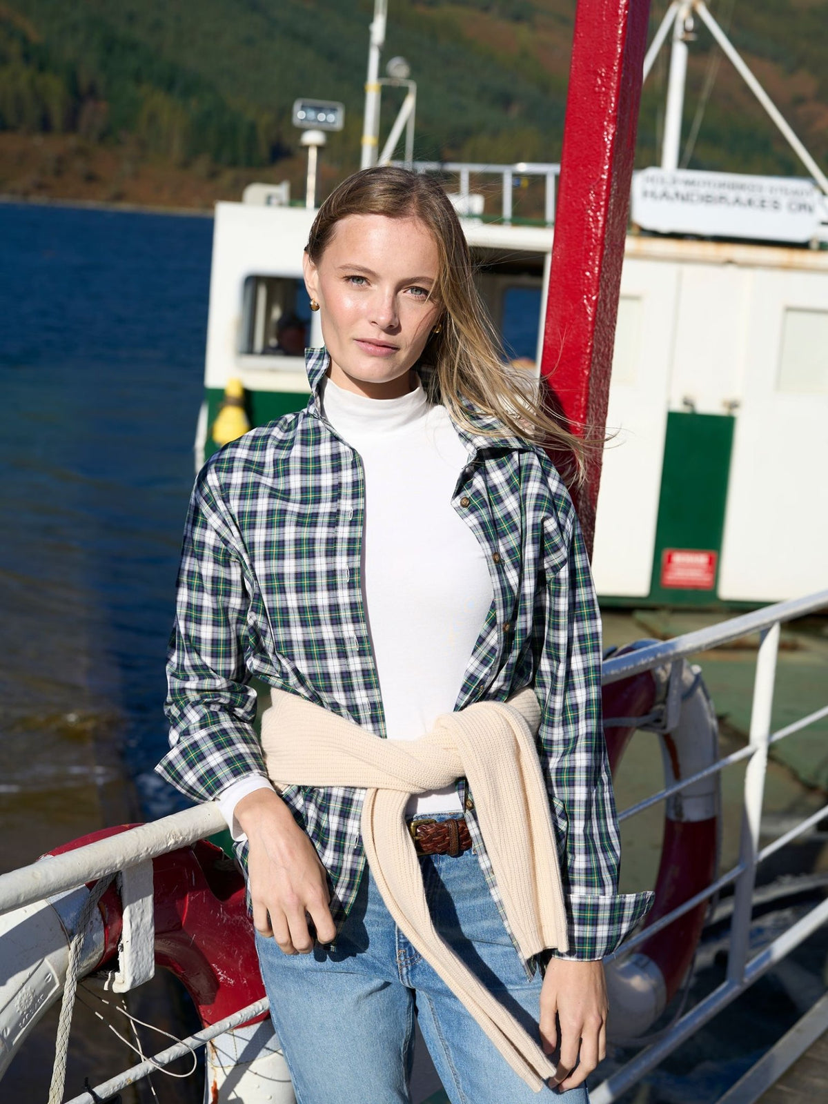 A woman wears a white turtleneck under a green and blue Cotton Tartan Shirt by Campbell’s of Beauly, with a cream sweater tied around her waist, while standing on the deck of a boat.