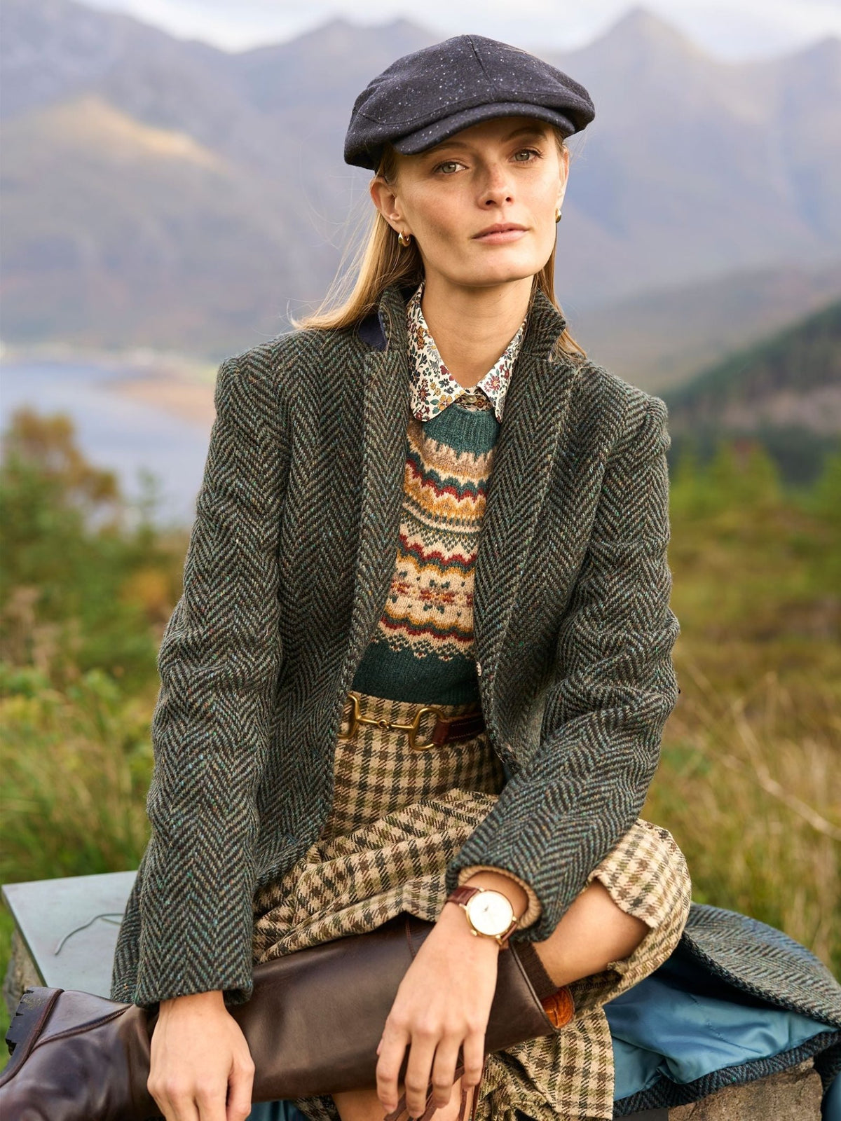 A person wearing a dark green herringbone Donegal Herringbone Coat by Campbell’s of Beauly over a patterned sweater, a plaid skirt, and a flat cap, sitting outdoors in a hilly landscape.