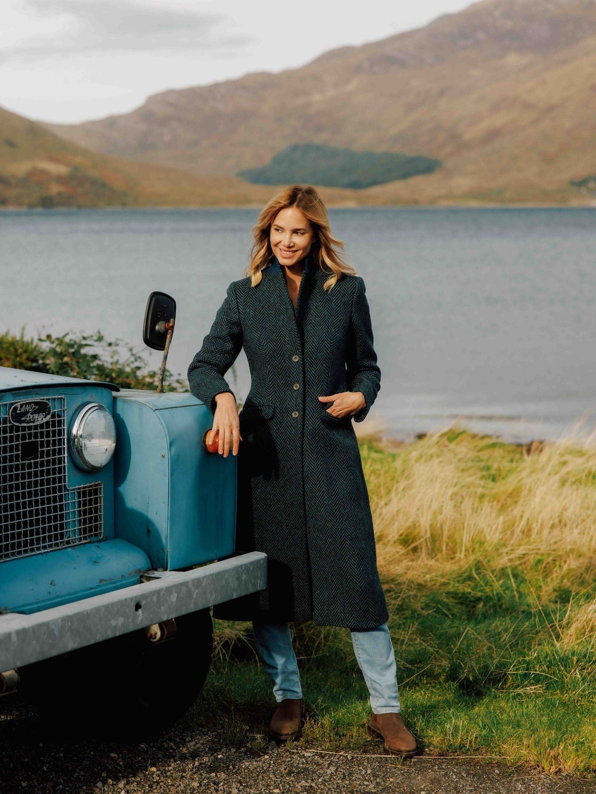 A woman wearing a long, dark herringbone Donegal Herringbone Coat by Campbell’s of Beauly leans against the front of a blue vintage Land Rover by a lake.