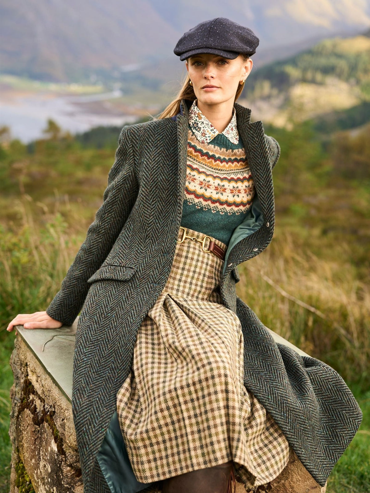A person wearing a dark green Campbell’s of Beauly Donegal Herringbone Coat over a patterned sweater and a plaid skirt, finished with a tweed flat cap.