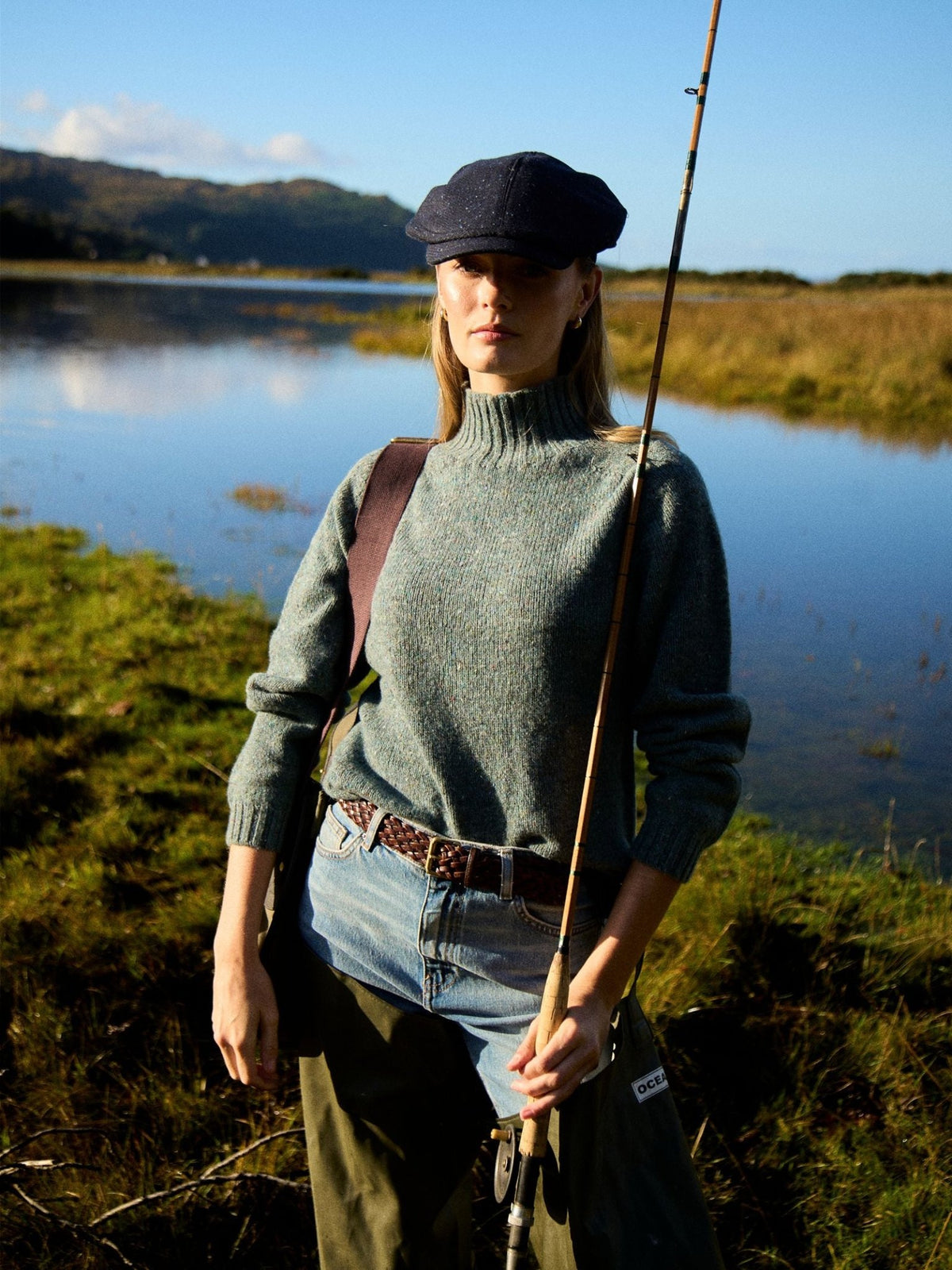 A woman wearing a grey Campbell’s of Beauly Donegal Lambswool Funnel Neck Jumper, flat cap, and waders, holding a fishing rod by a lake.