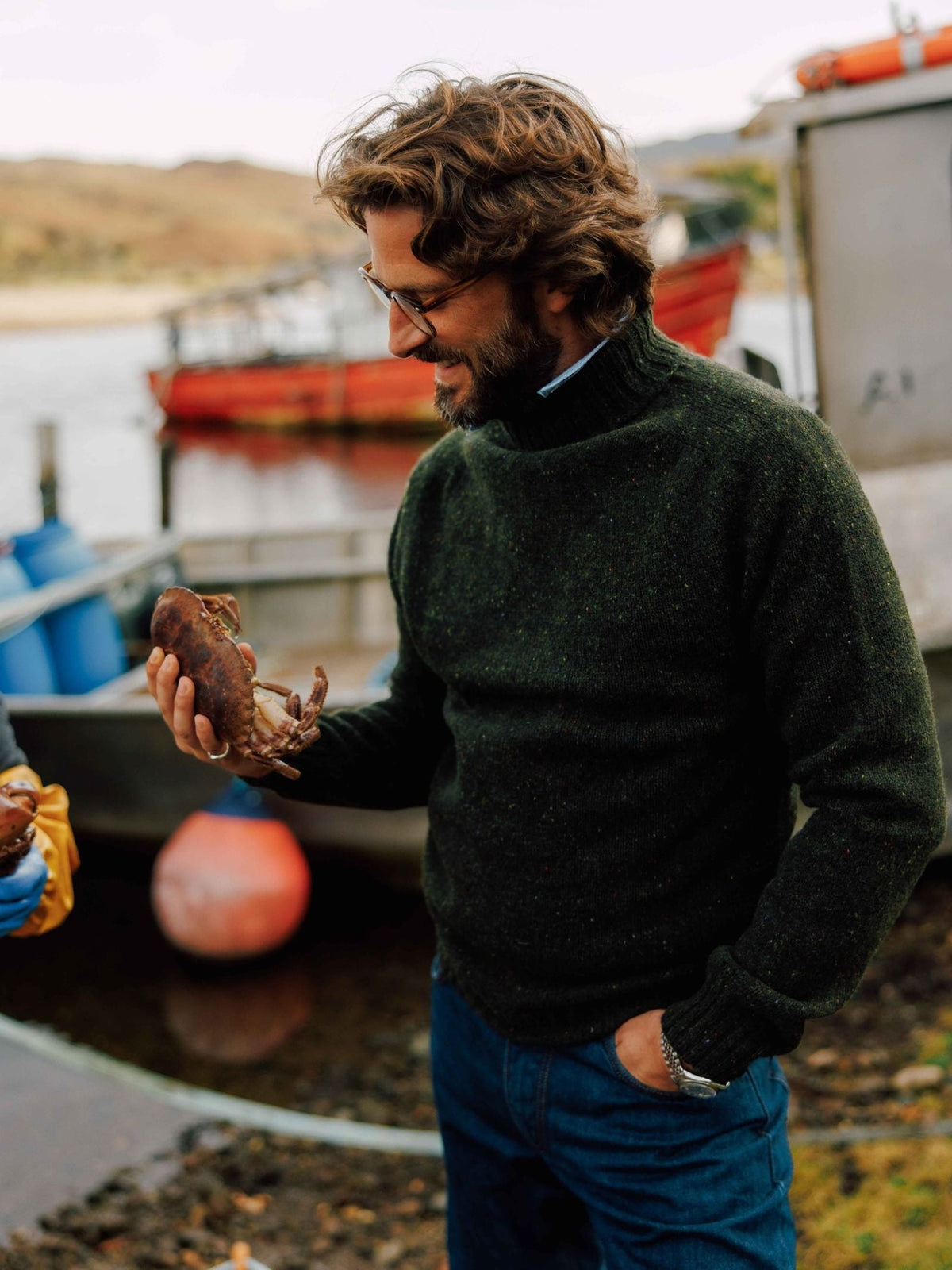 A man wearing a dark green Donegal Lambswool Roll Neck sweater smiles while holding a crab by a boat at a harbor.