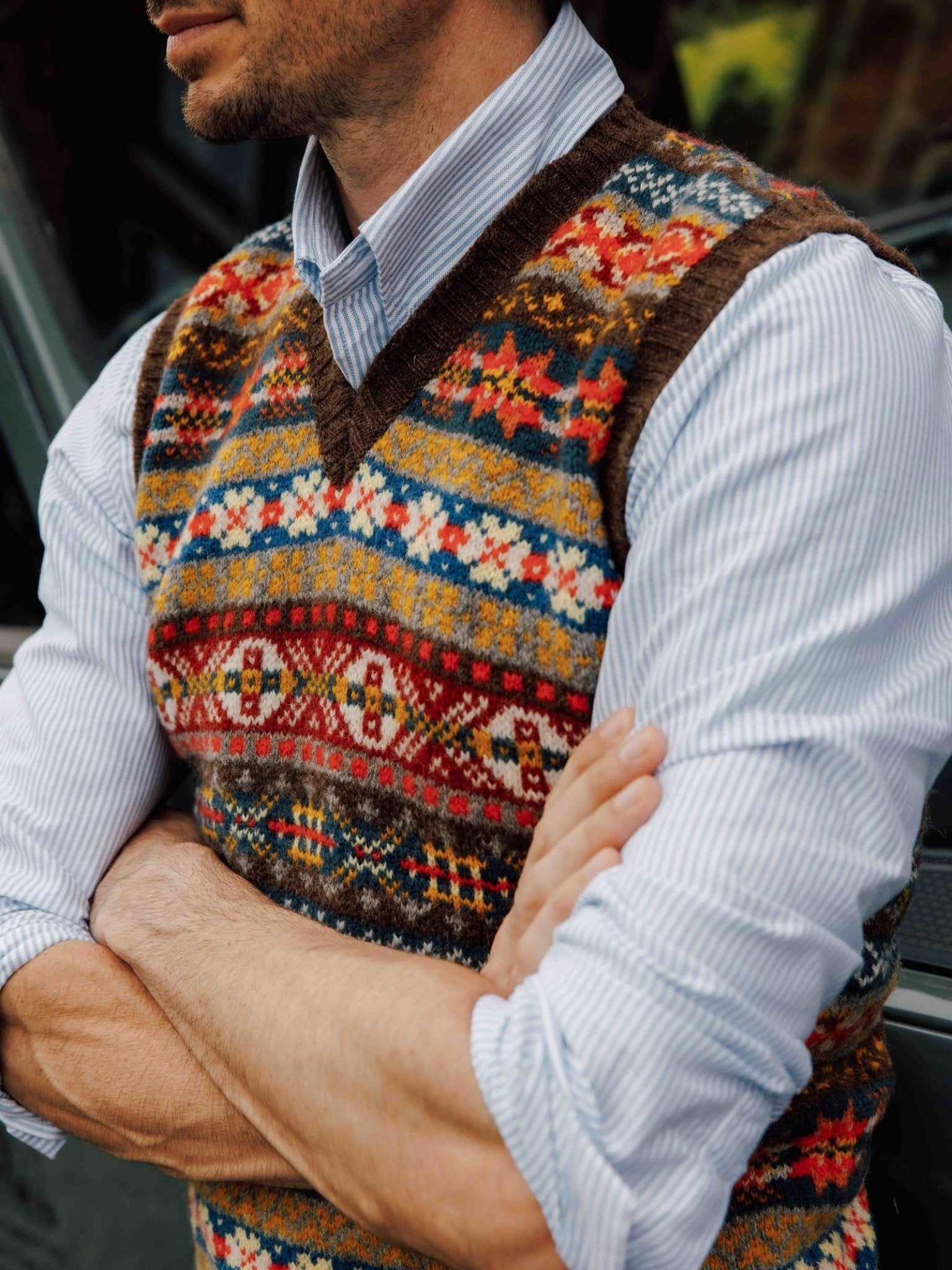 A person with arms crossed wears a blue striped shirt and the Campbell’s of Beauly Fairisle Slipover—a sleeveless knit layer crafted from soft Shetland wool, featuring colorful geometric patterns.