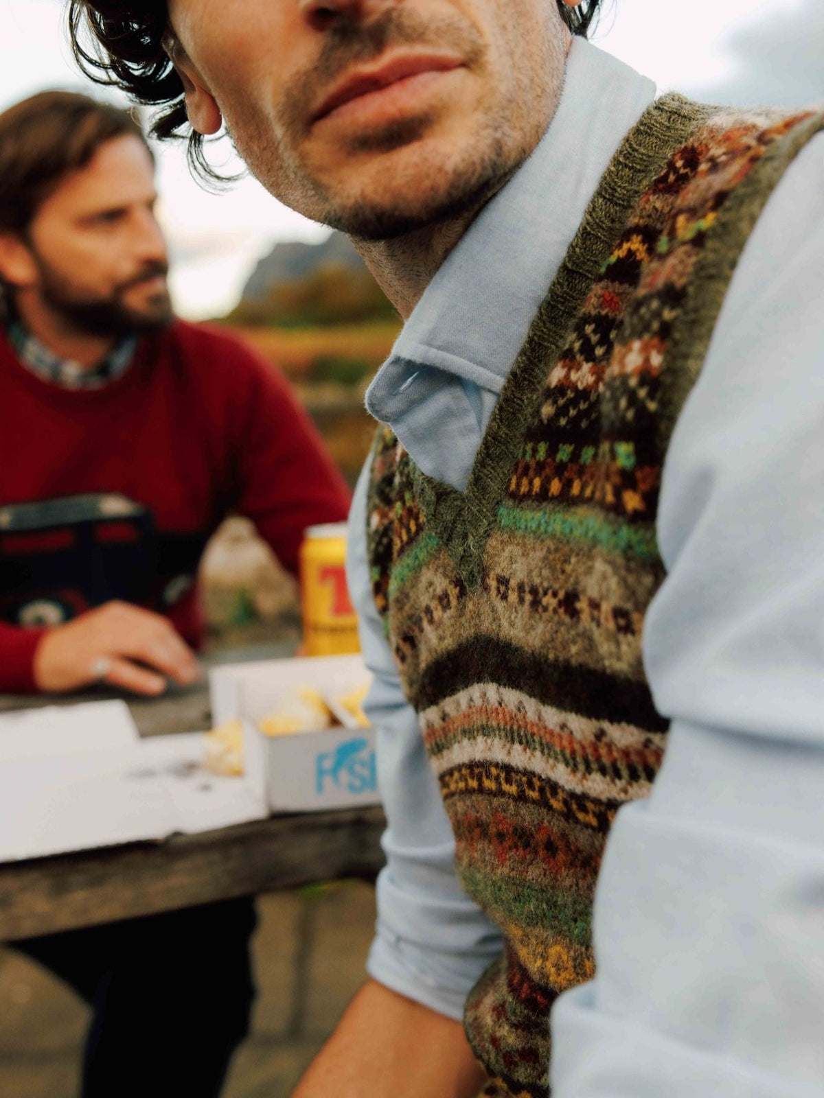 Two men sit at an outdoor table with a box of food and a drink. The man in front wears the Campbell’s of Beauly Fairisle Slipover over a blue shirt, creating a casual and relaxed scene.