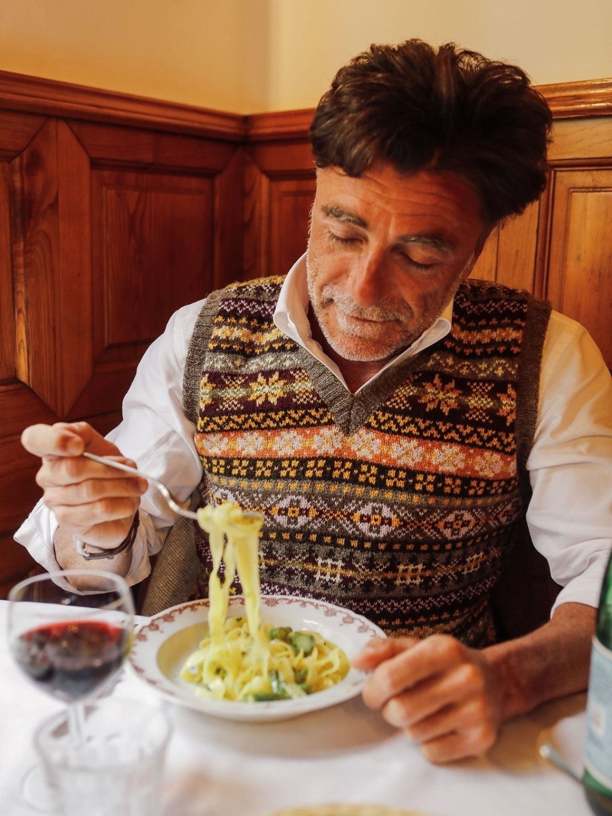 A man in the Campbell’s of Beauly Fairisle Slipover—a timeless sleeveless knit made from soft Shetland wool—sits at a table in a cozy restaurant, looking down at his pasta while a glass of red wine and bottled water rest nearby.
