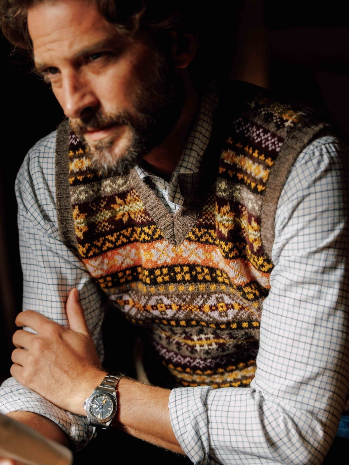 A bearded man wears a Campbell’s of Beauly Fairisle Slipover layered over a light-colored checkered dress shirt.