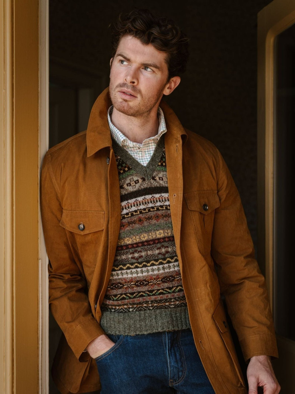 A man with curly hair stands in a doorway, one hand tucked into the pocket of his jeans, gazing thoughtfully to the side. He wears a brown jacket layered over a Campbell’s of Beauly Fairisle Slipover and a collared shirt.