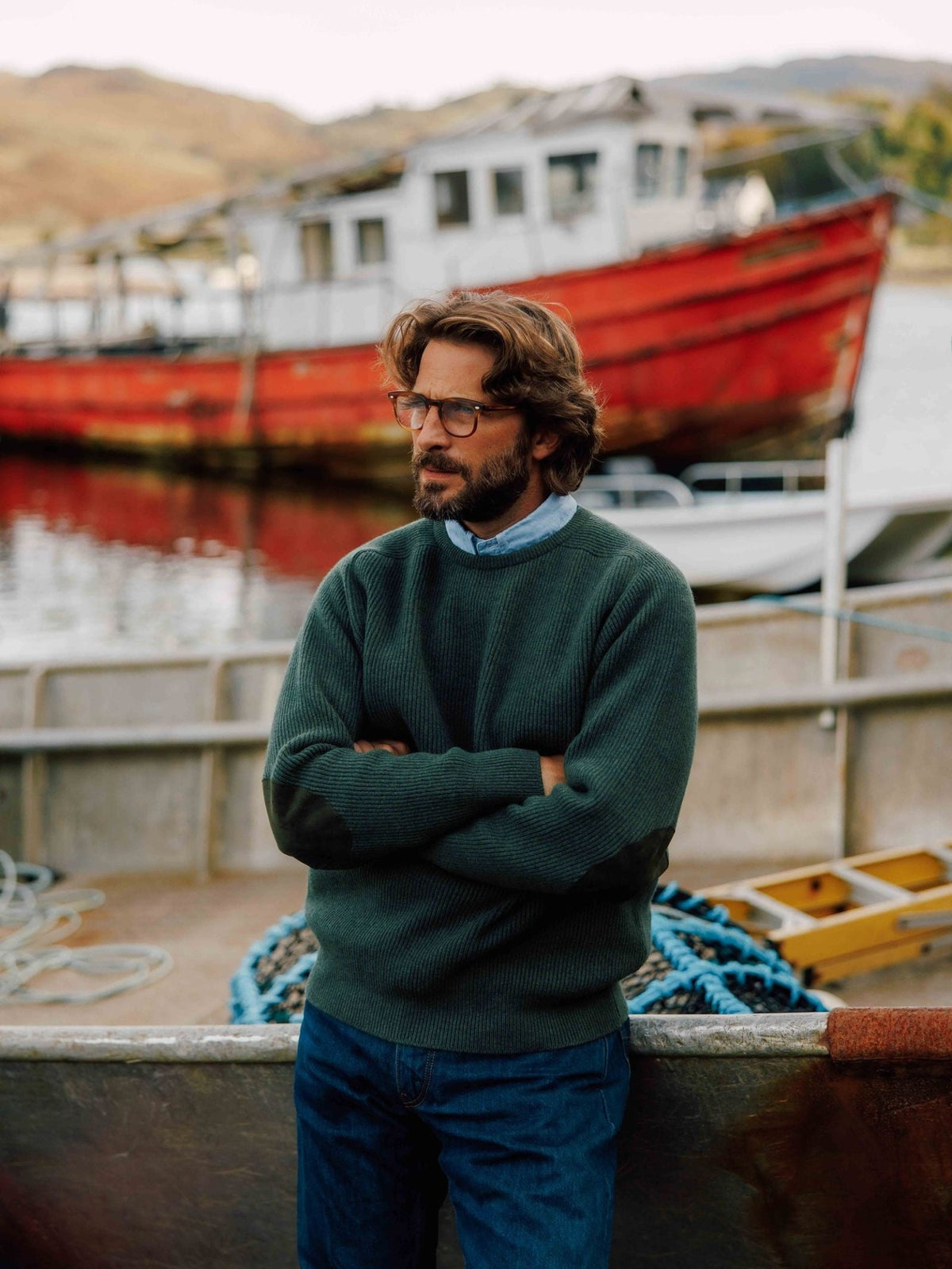 A bearded man wearing glasses and a green Fisherman’s Rib Crew Jumper by Campbell’s of Beauly with suede elbow patches, standing on a boat dock in front of a red fishing vessel.