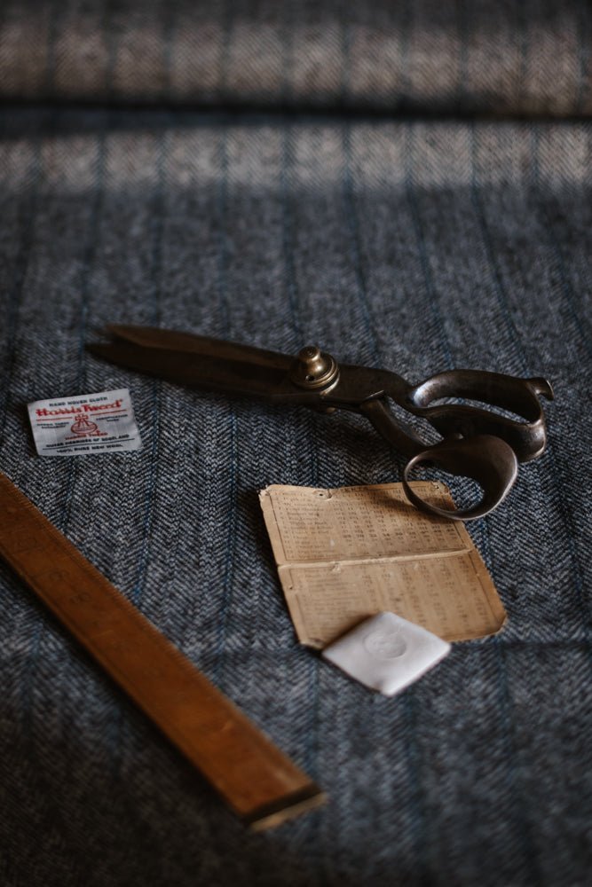 Large metal scissors, a wooden ruler, a small packet of needles, an old piece of folded paper, and a white square cloth rest on the textured grey herringbone House Tweed 95053 fabric by Campbell’s of Beauly.