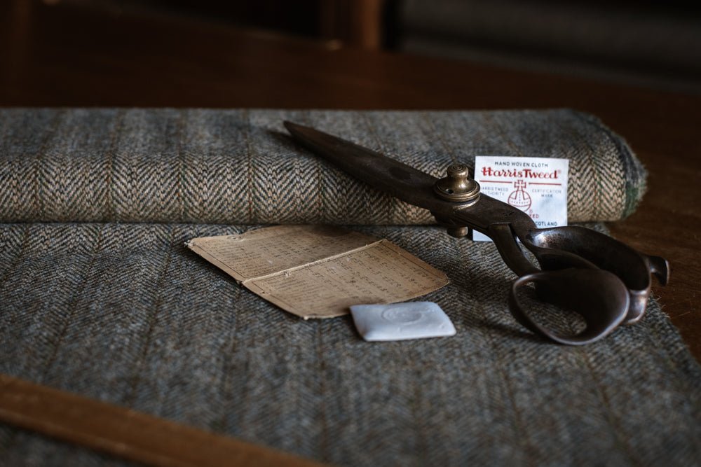 House Tweed 95054 by Campbell’s of Beauly, paired with large vintage scissors, a handwritten note, a piece of chalk, a wooden ruler, and a Harris Tweed label set upon a wooden table, evokes the timeless tradition of bespoke tailoring. The classic herringbone pattern of this jacketing cloth further highlights the enduring craftsmanship associated with House Tweed 95054.