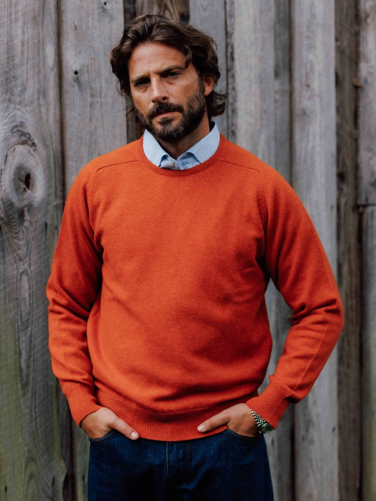 A man wearing a bright orange Campbell’s of Beauly Lambswool Crew Neck Jumper layered over a light blue collared shirt.