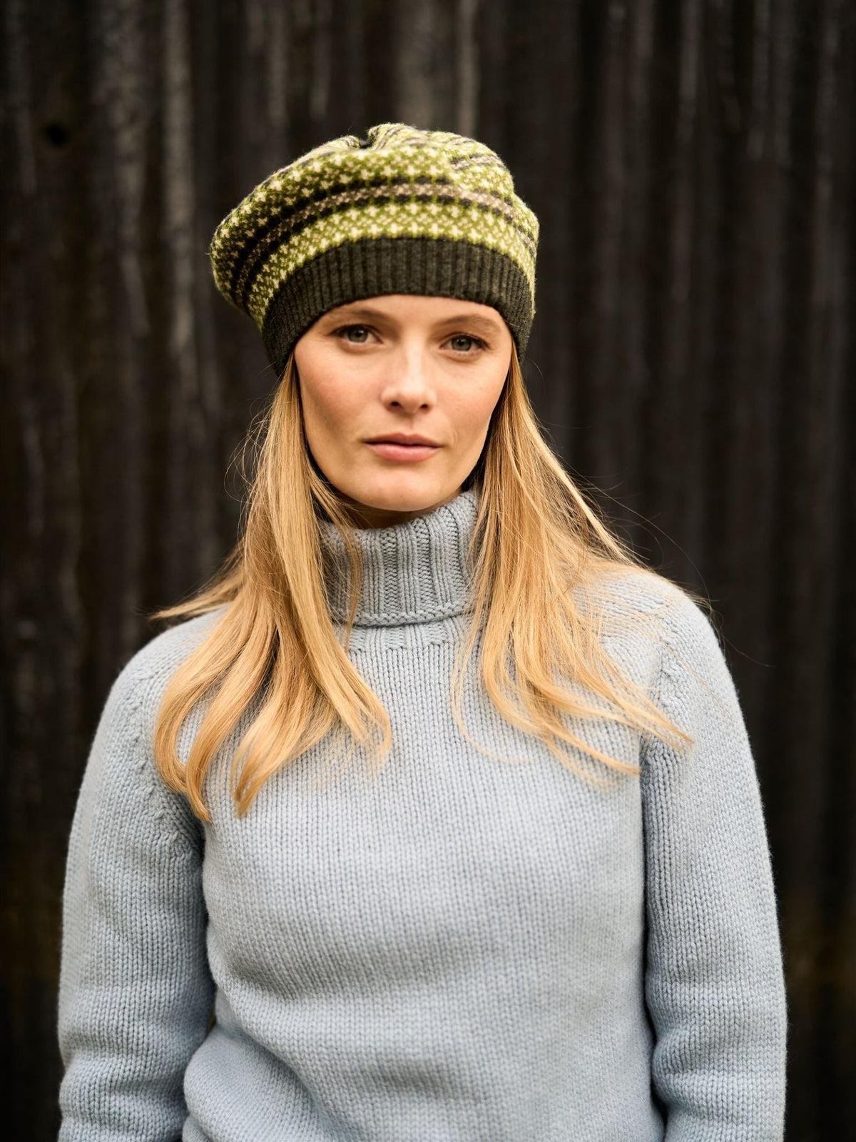 A woman wears a green and olive Lambswool Fairisle Beret by Campbell’s of Beauly with a light blue turtleneck sweater.