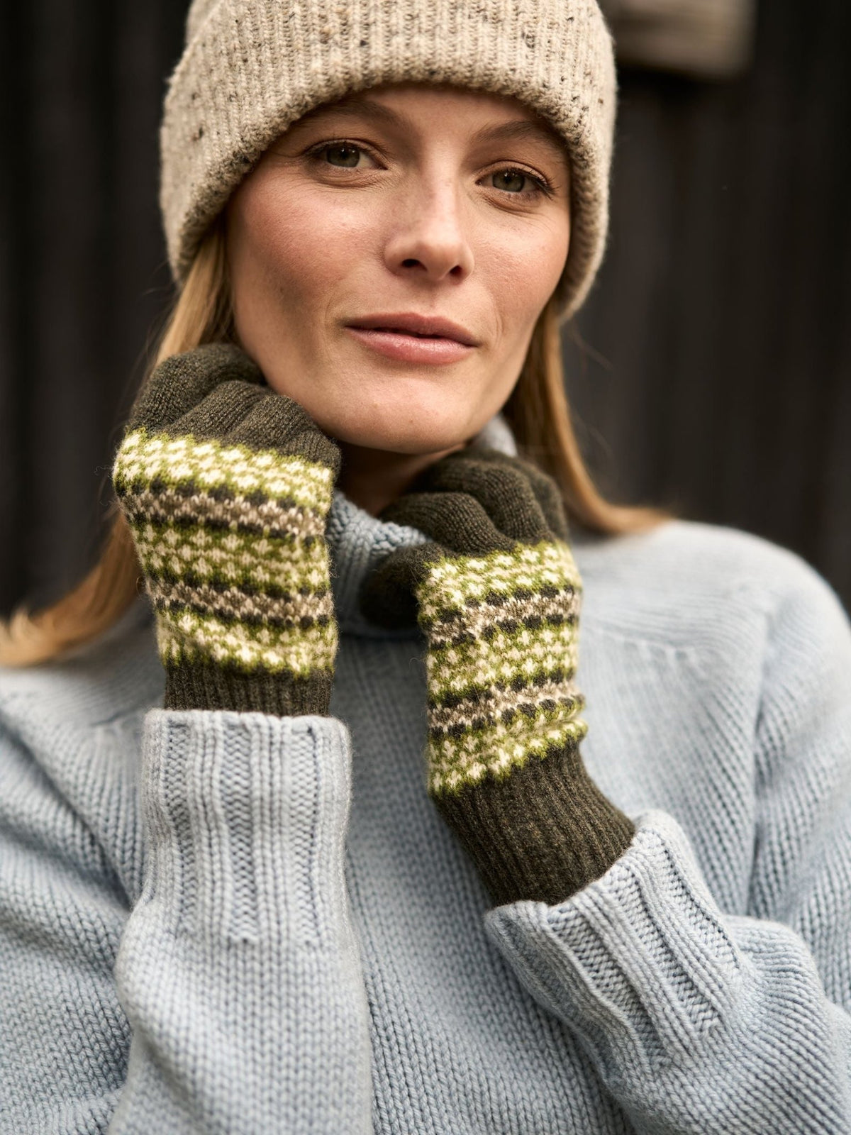 A woman wearing a light blue sweater and a knit beanie holds her hands up to display olive green Lambswool Fairisle Gloves featuring a multicolored Fair Isle pattern.