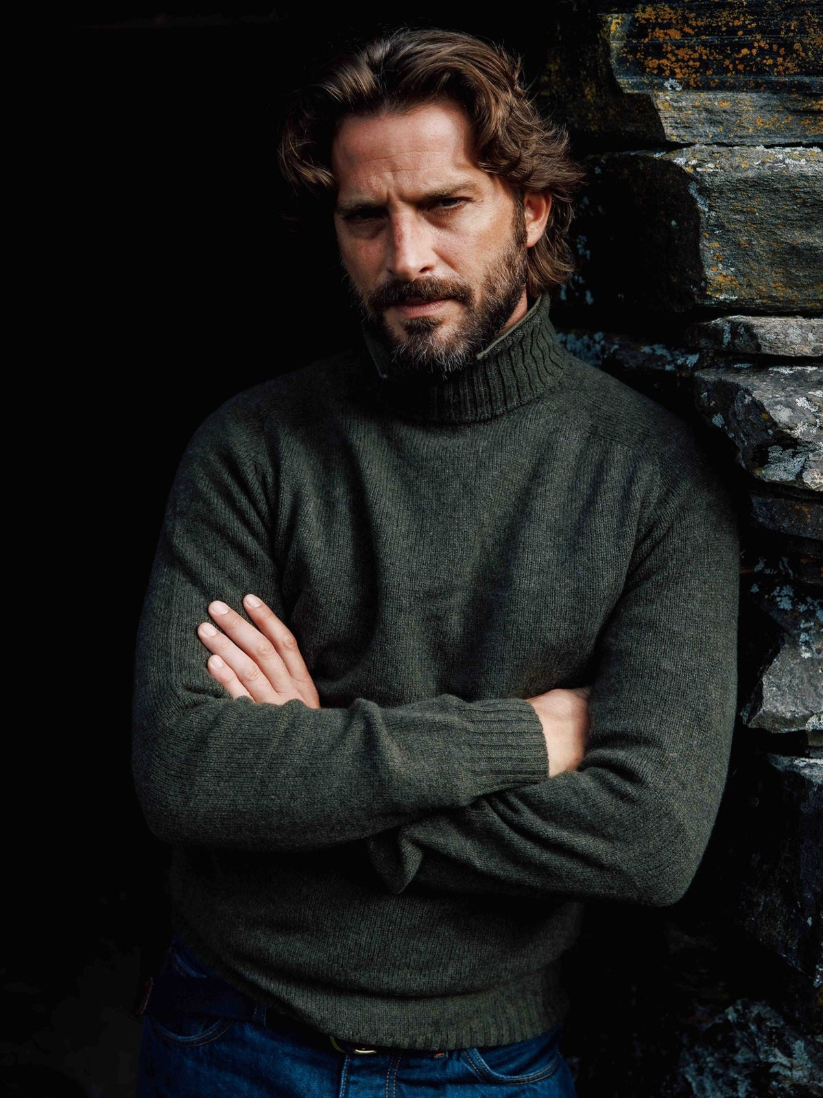 A bearded man with his arms crossed, wearing an olive green Campbell’s of Beauly Lambswool Roll Neck.