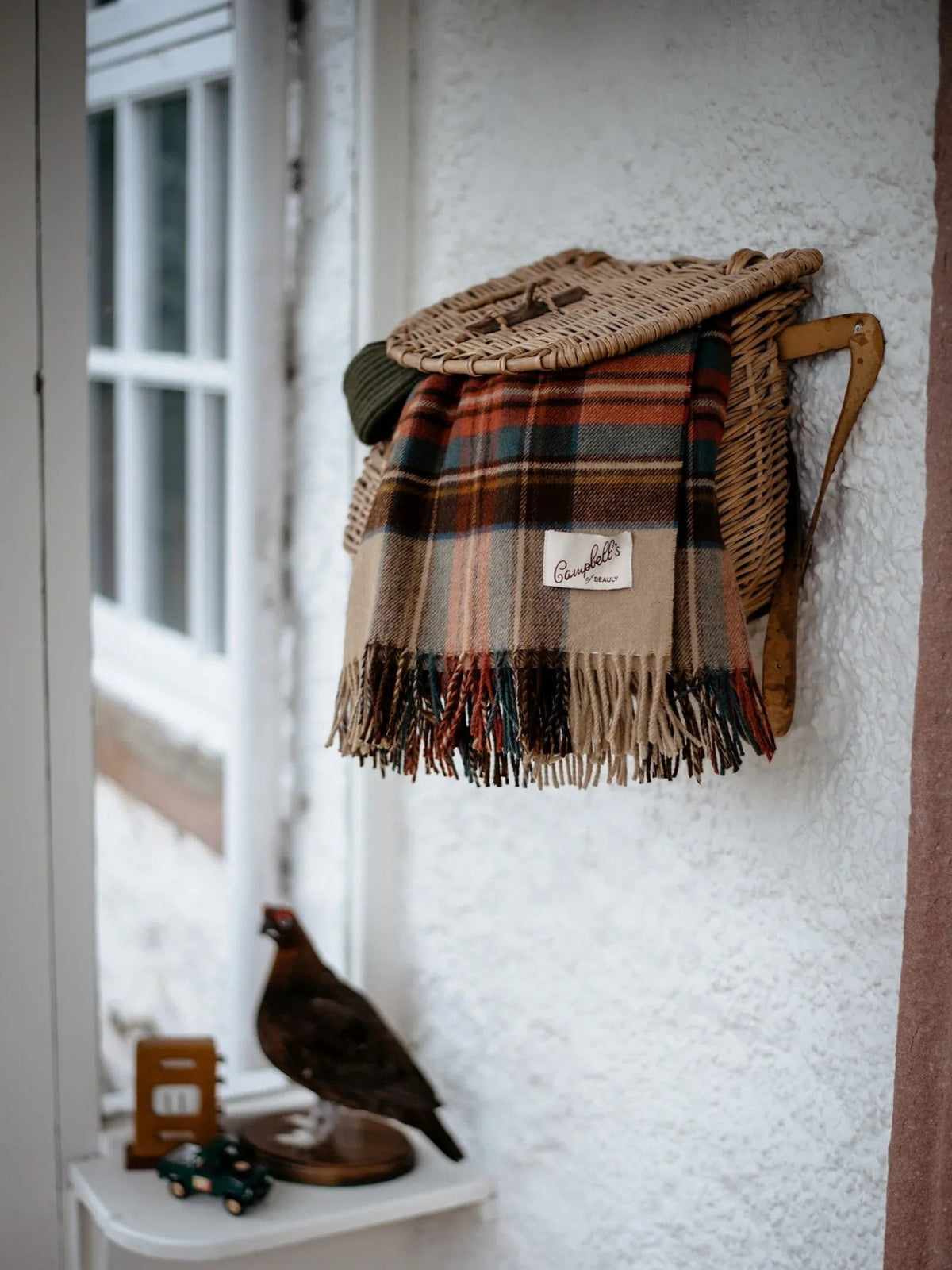 A Lambswool Tartan Blanket by Campbell’s of Beauly drapes elegantly over a wicker rack mounted on a white wall, introducing heritage charm to the space. Below, a decorative bird figure and small wooden items are arranged on a narrow shelf near the window.