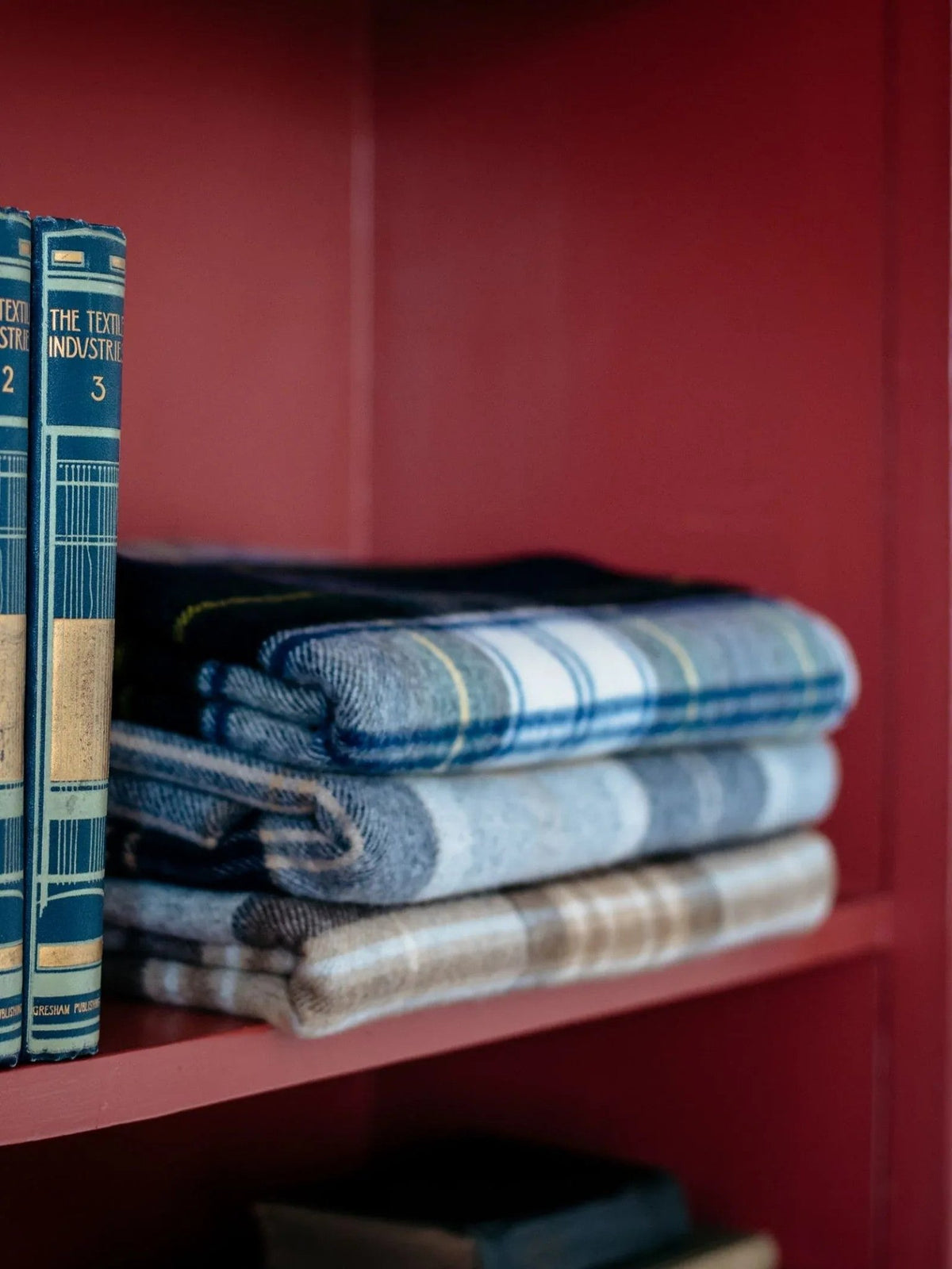 Three neatly folded Campbell’s of Beauly Lambswool Tartan Blankets sit on a wooden shelf next to three vintage blue books, all within a red bookcase, bringing heritage charm and warmth to the space.