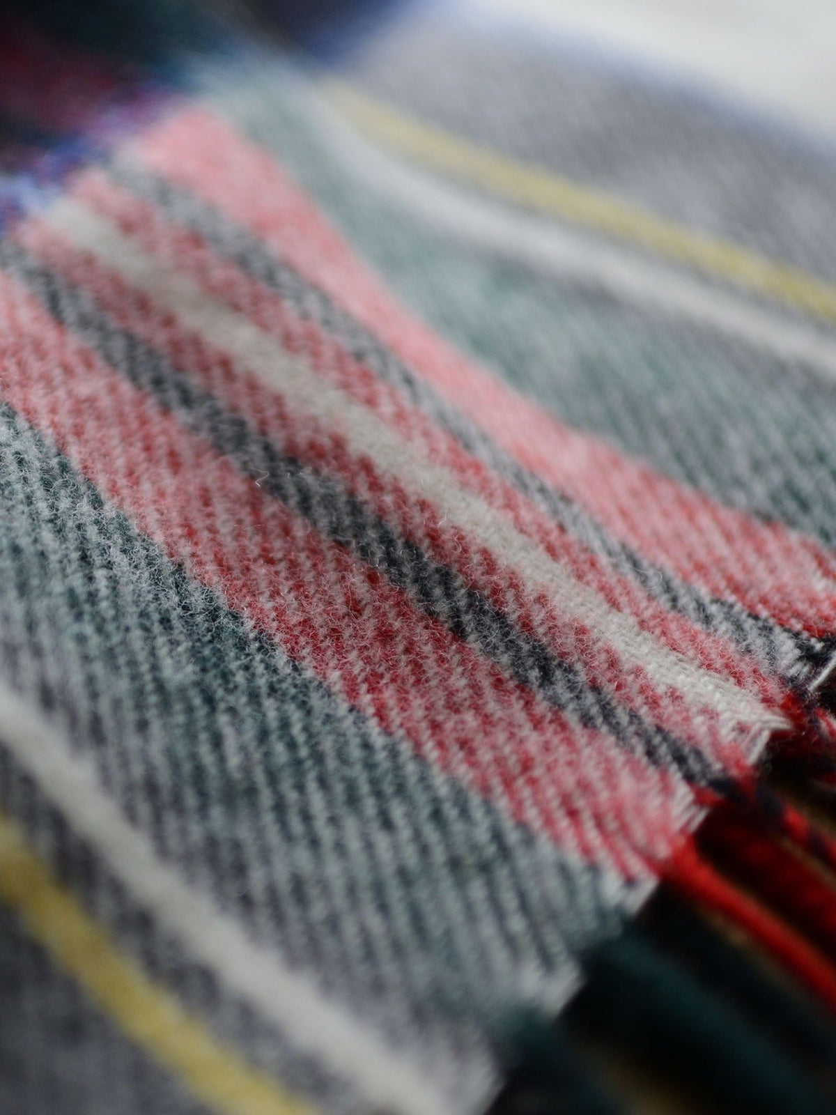 Close-up of a soft, woven lambswool fabric showcasing a plaid pattern in red, gray, white, and subtle yellow accents. The fine texture and fringed edge highlight the heritage charm of the Campbell’s of Beauly Lambswool Tartan Blanket.
