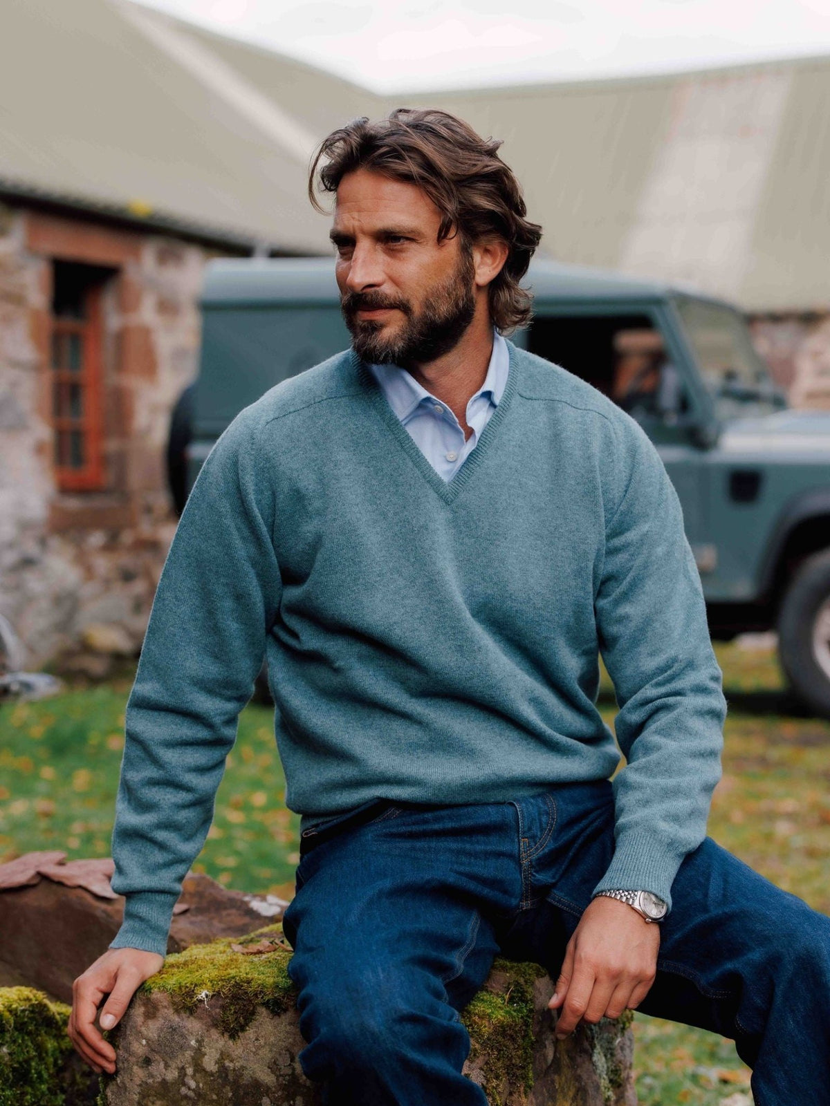 A man wearing a Campbell’s of Beauly Lambswool V-Neck Jumper over a light blue collared shirt and jeans sits on a mossy stone wall in front of a green vehicle.