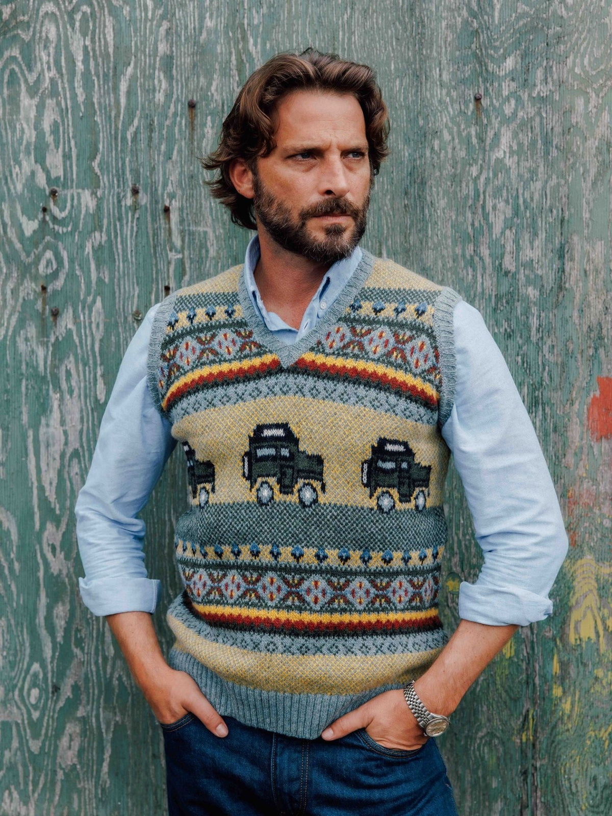 A man wearing a Land Rover Defender Fairisle Slipover patterned with off-road vehicles over a light blue button-down shirt.
