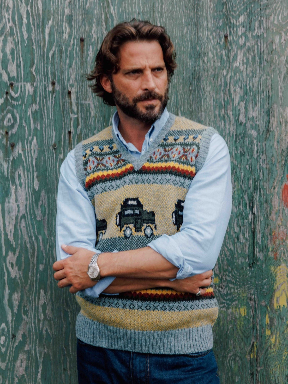 A man wears a light blue button-down shirt layered under a Land Rover Defender Fairisle Slipover in premium wool with a repeating off-road vehicle motif.