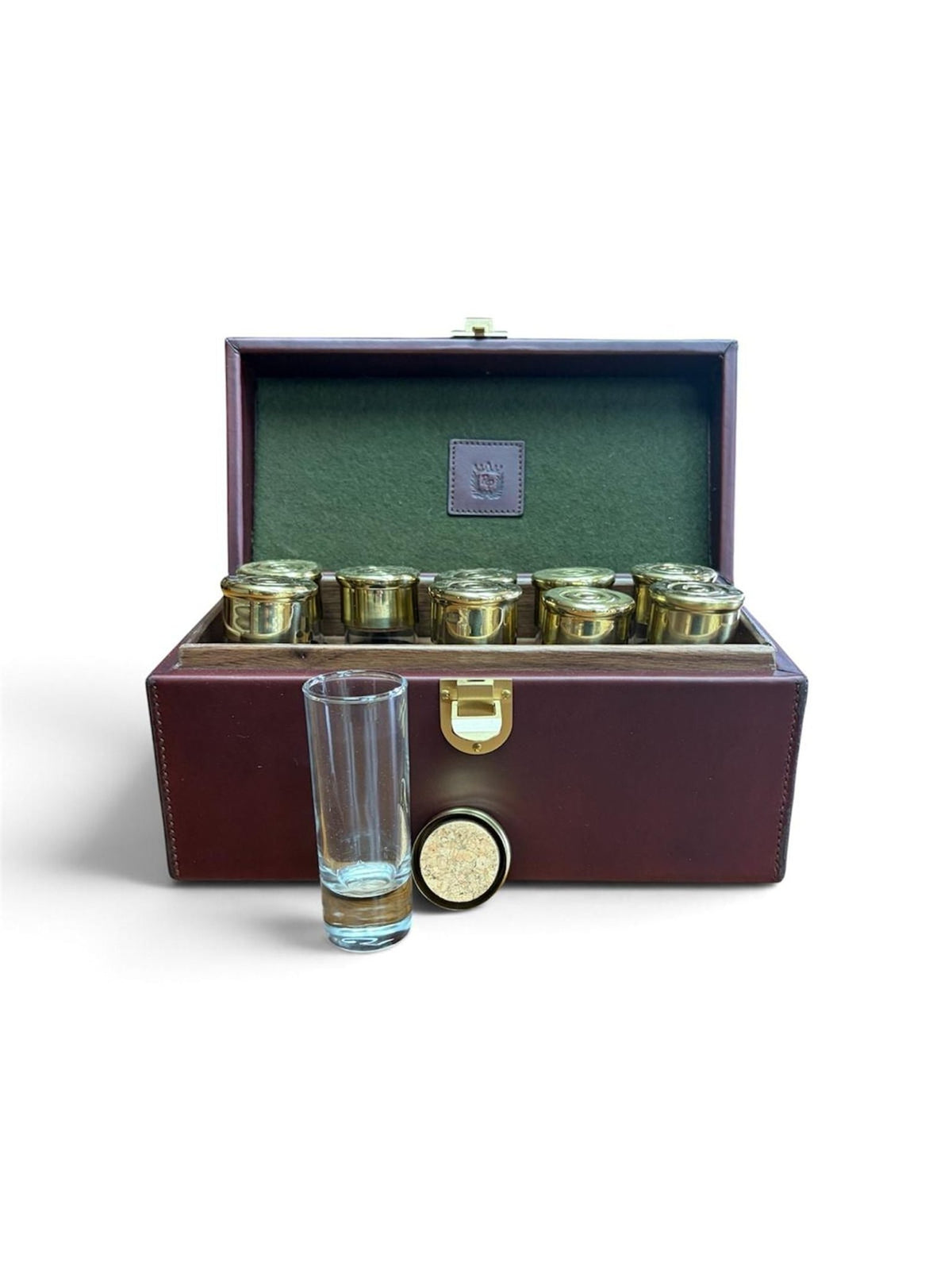 The Rey Pavón Leather Cartridge Shot Glass Set features a brown leather case with brass accents, holding six cartridge-shaped shot glasses inside. In front of the set, a clear empty glass and a round cork lid rest on the white surface.