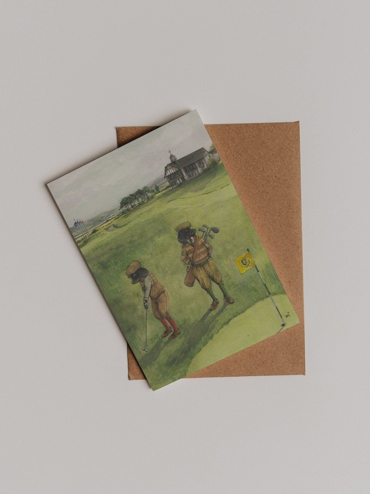 A Links Dornoch Greeting Card from Campbell’s of Beauly, featuring an illustration of two children golfing on green hills, is partially tucked into a brown envelope and displayed on a plain background.