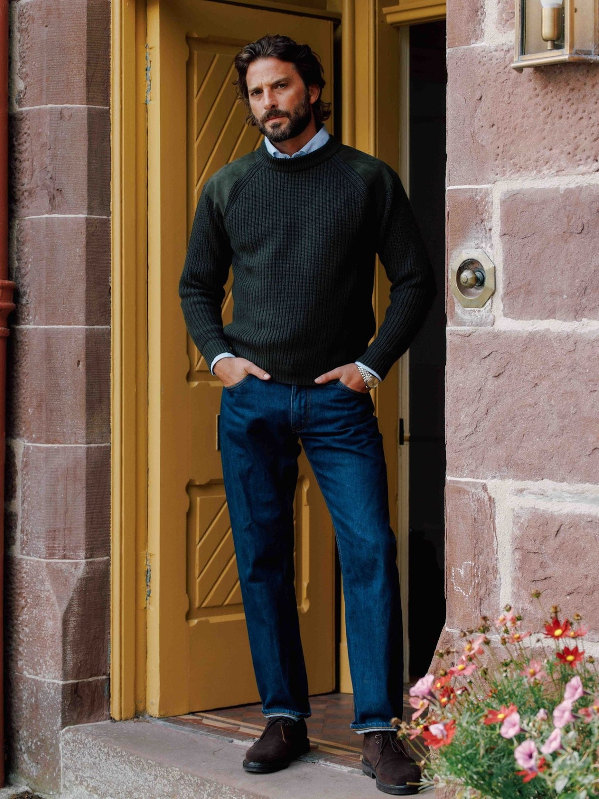 A man wearing a Campbell’s of Beauly Lux Cashmere Shooter Crew Jumper with shoulder patches, a light blue collared shirt, and dark blue jeans standing in a doorway.